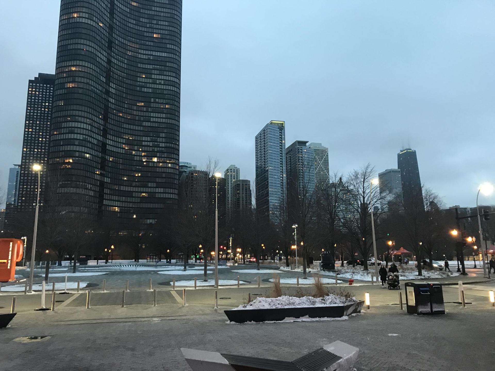 A city park with bare trees and snow, surrounded by tall buildings at dusk.