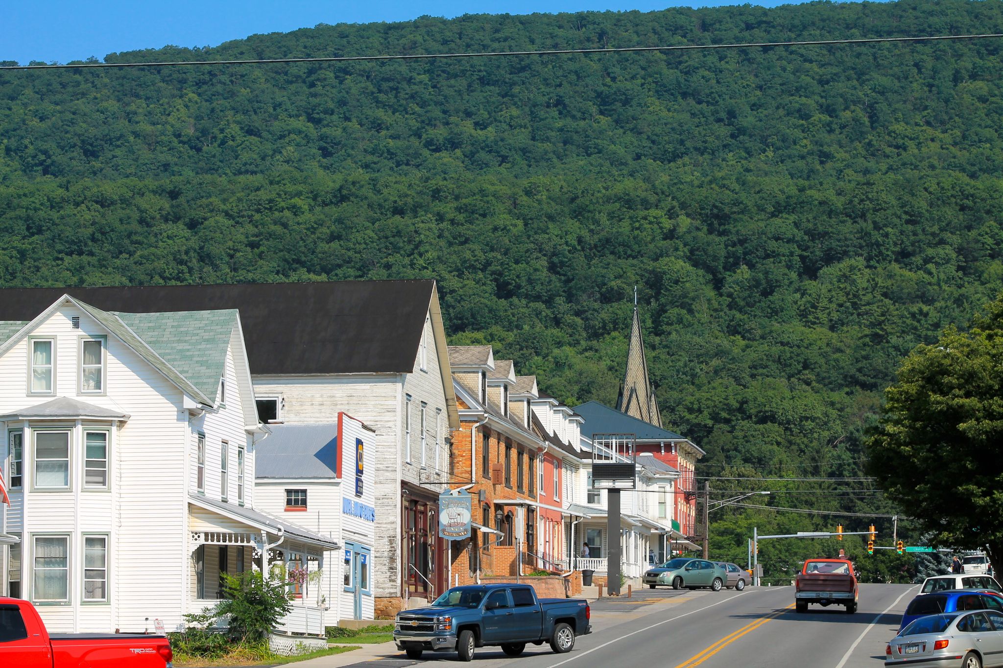 Sunny street with buildings, cars, and a steep, forested hillside. - Centre Hall to Millheim loop