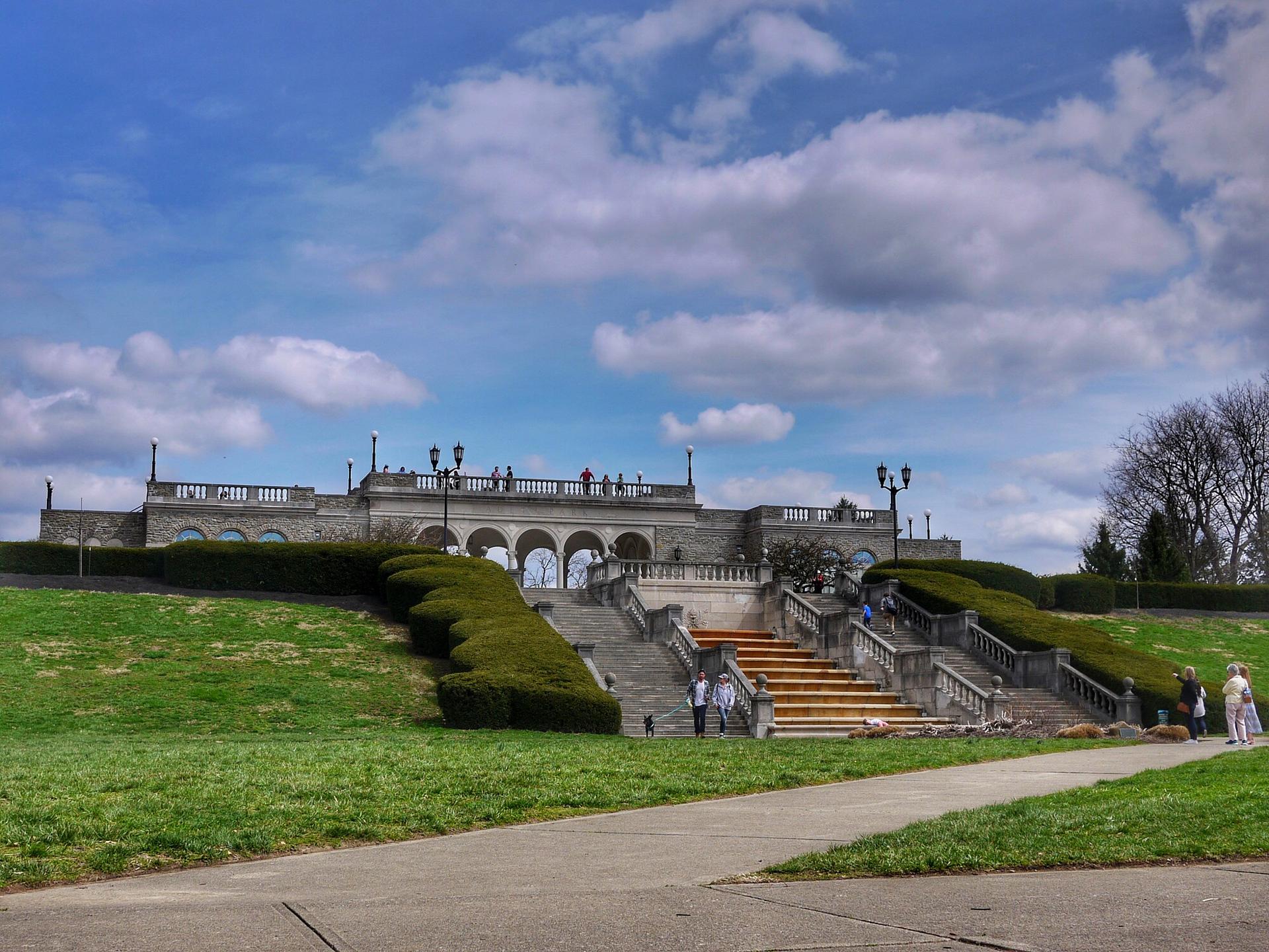 Grand stone building with terraces and stairs on a grassy hill, people walking.