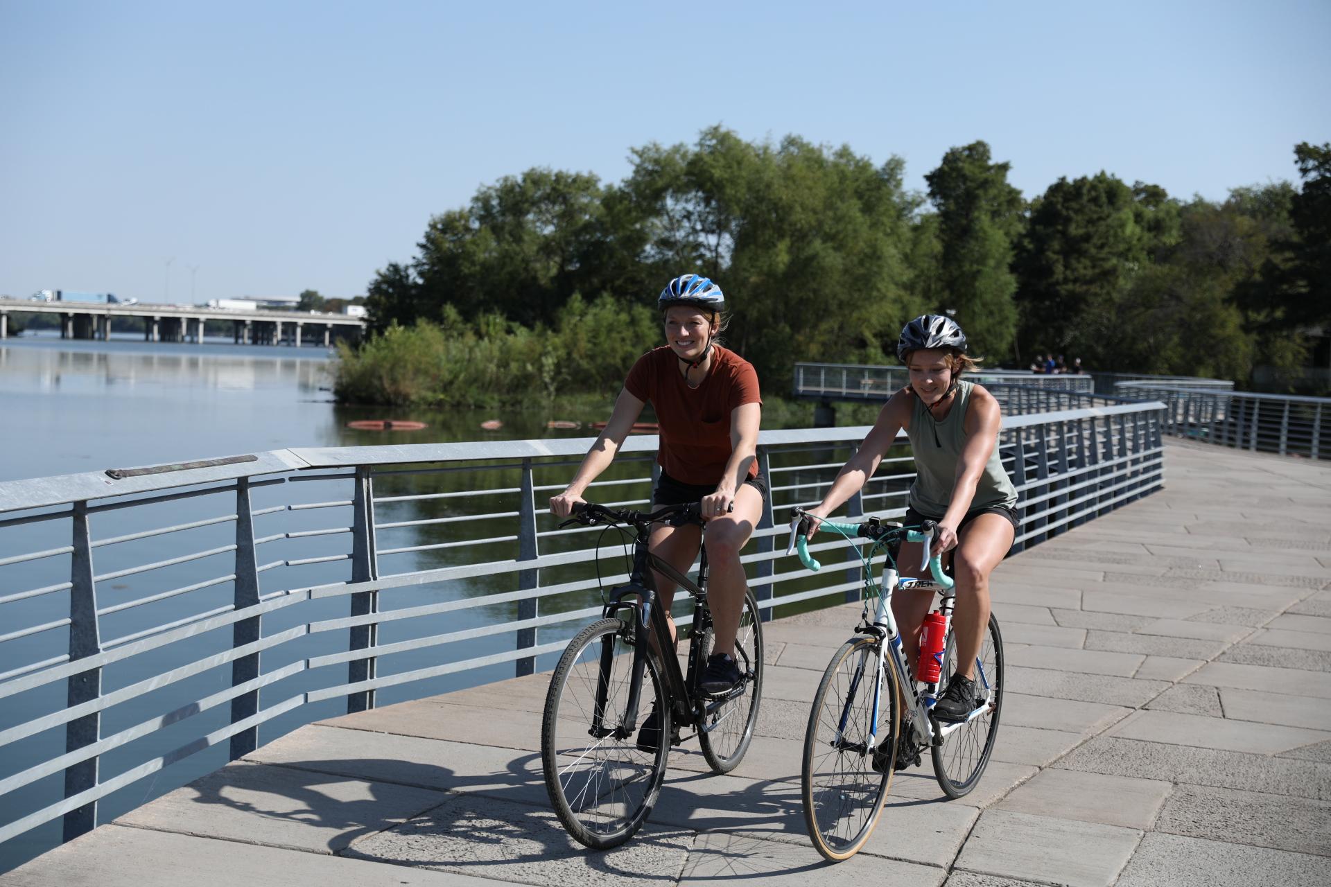 Two people cycling on a bridge next to a river on a sunny day.