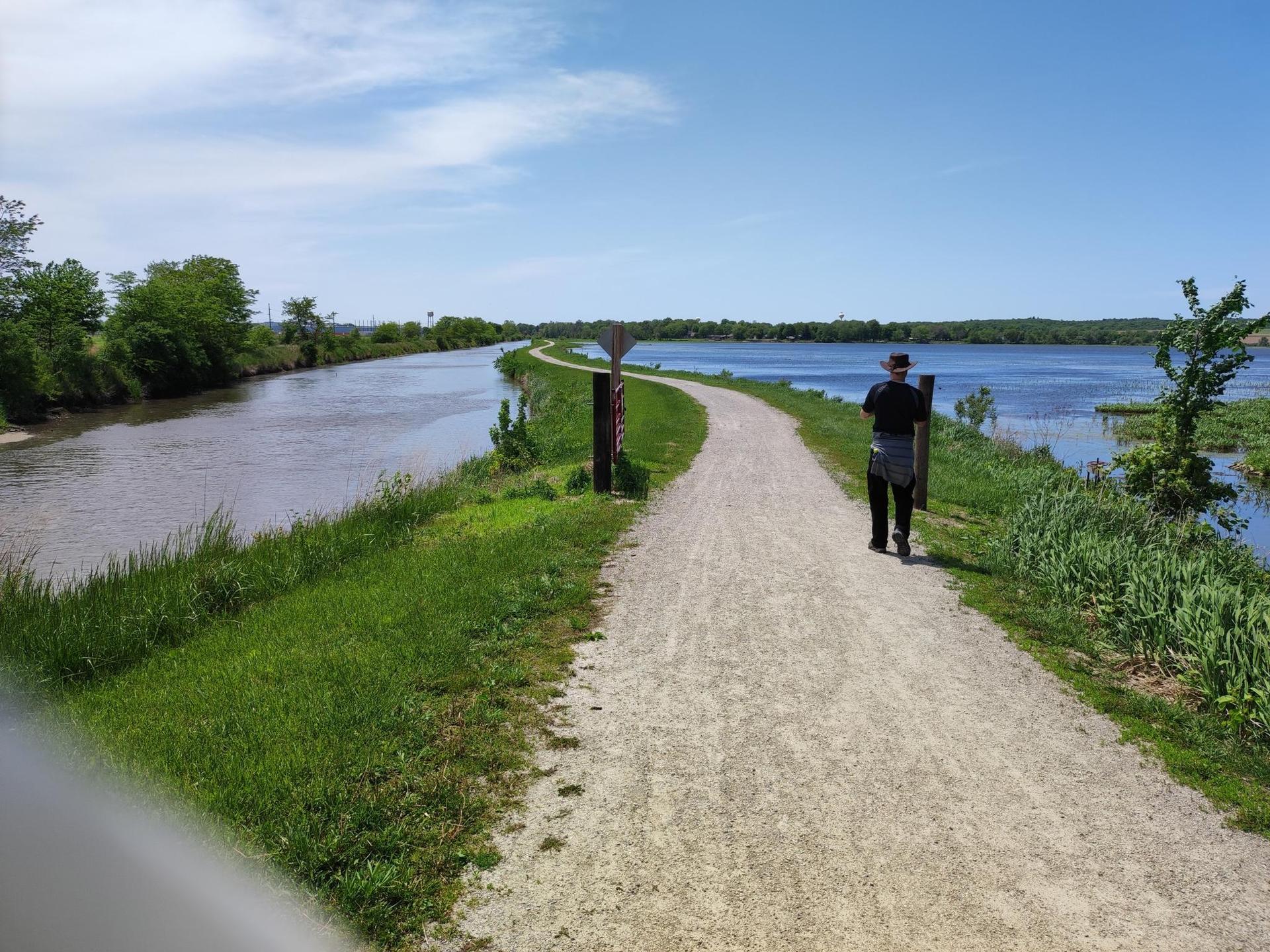 Person walks on a sunny gravel path between two waterways with green banks.