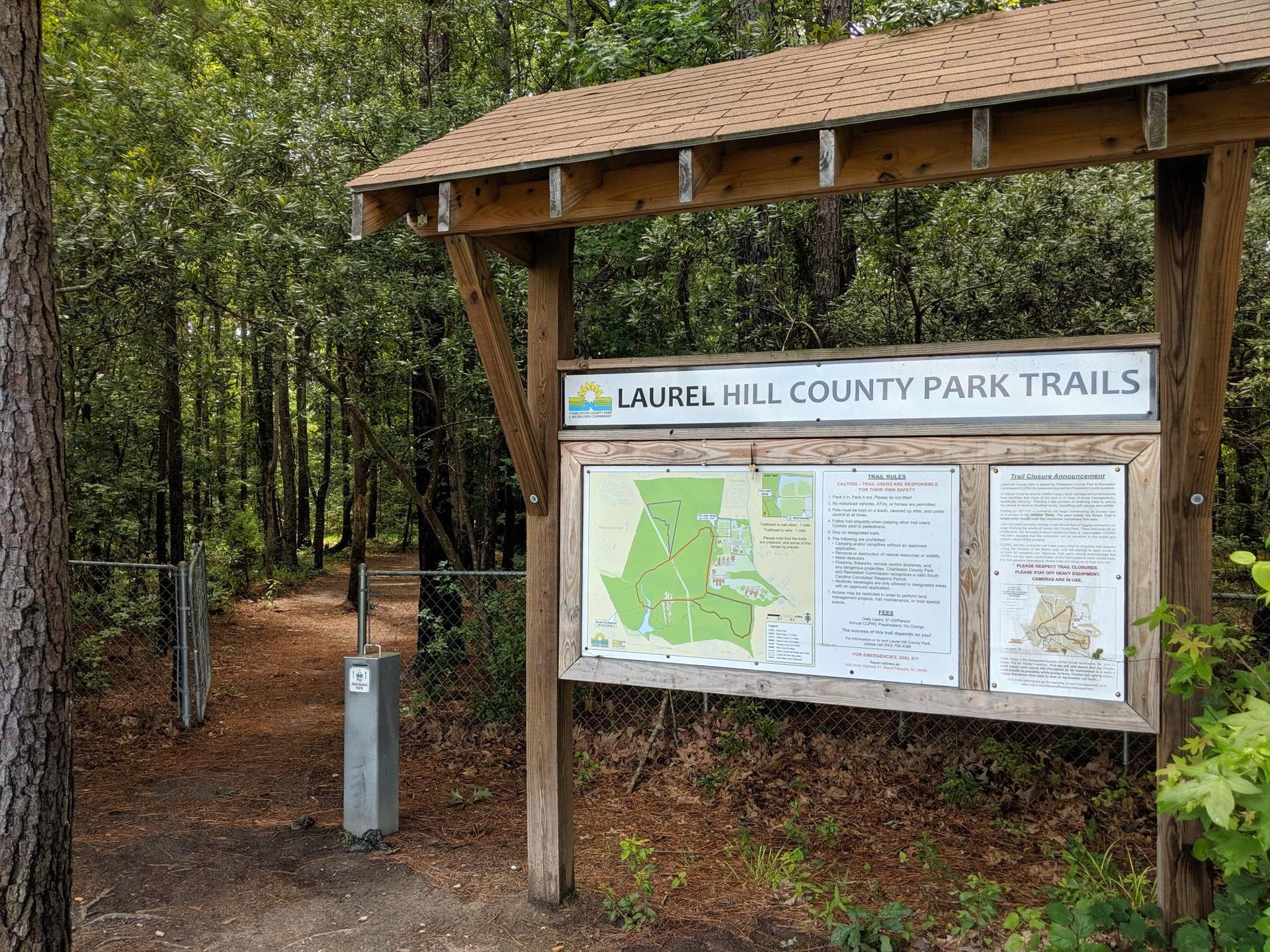 Wooden information board with maps beside a dirt path entering a forest.