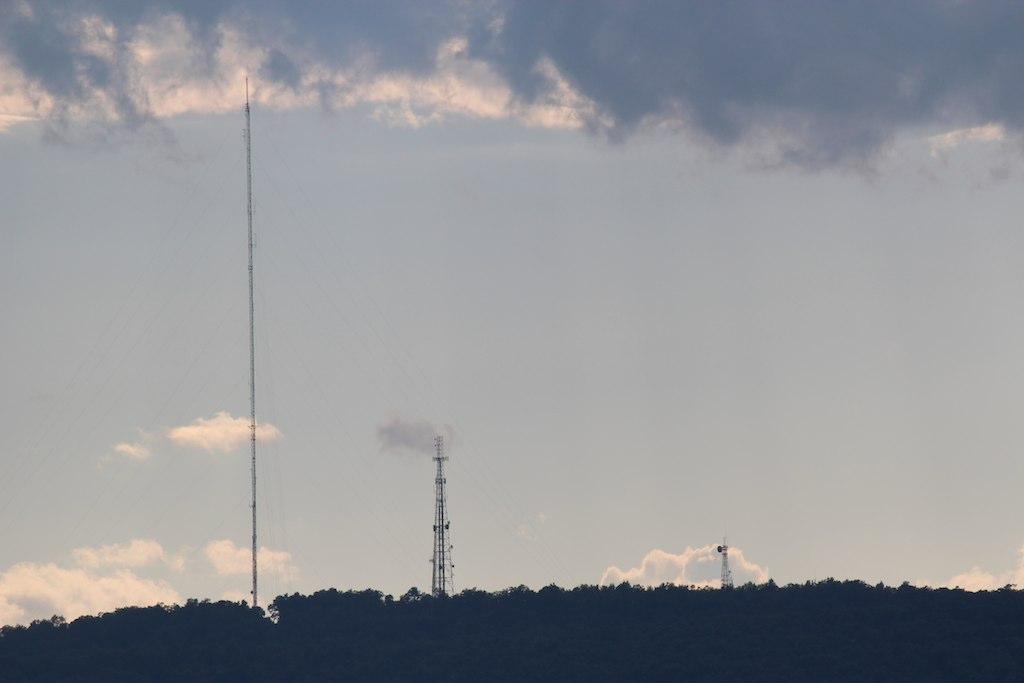 Three communication towers silhouetted against a cloudy sky above a hill.
