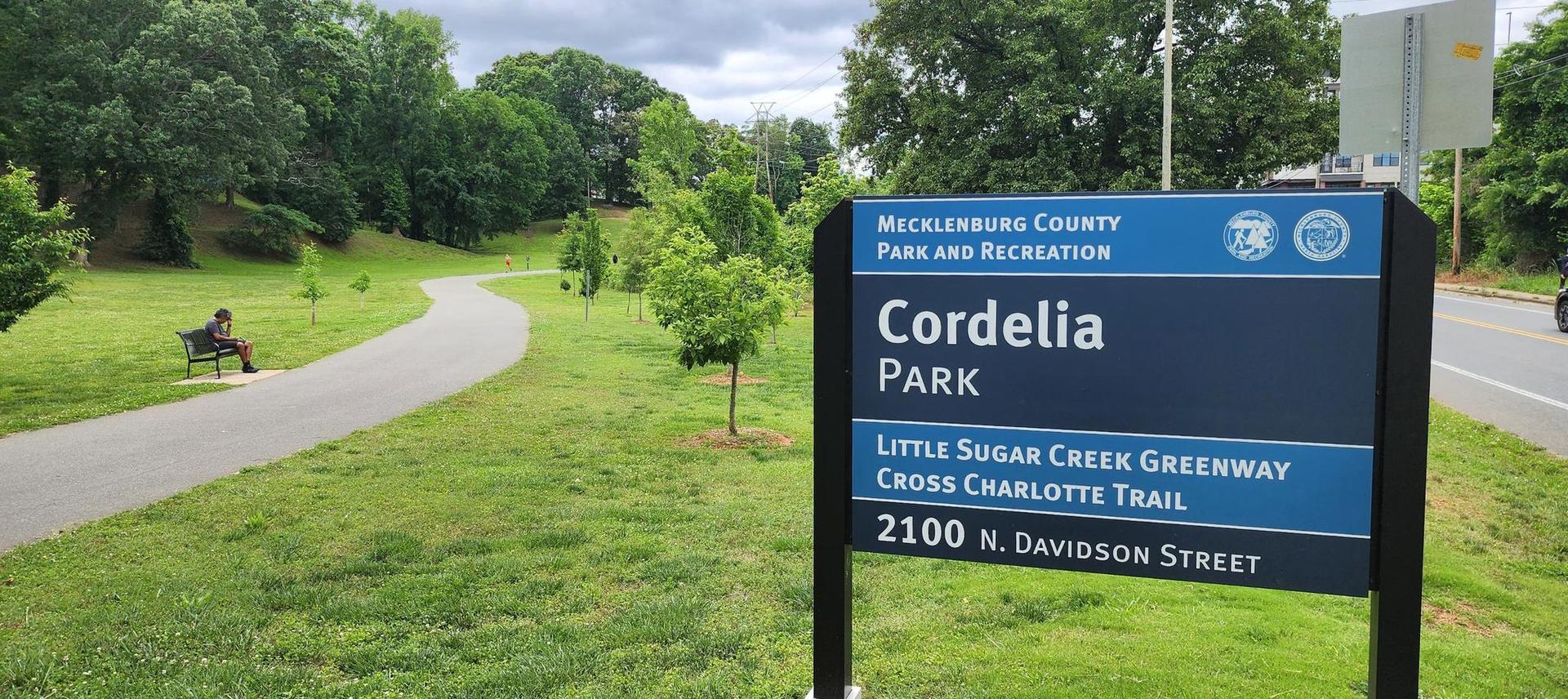 Overcast park scene with a sign, paved path, and person on a bench.