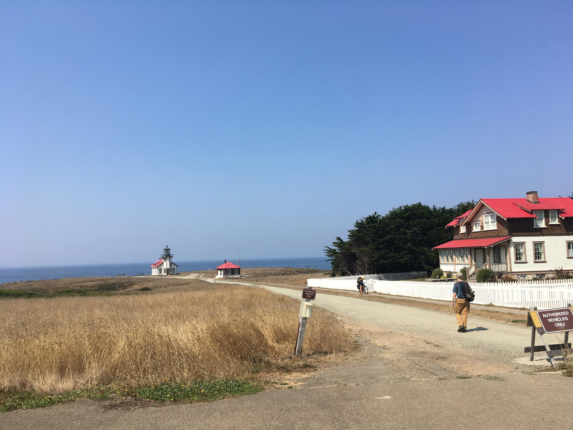 Lighthouse and red-roofed buildings by the ocean under a clear sky, person walking.