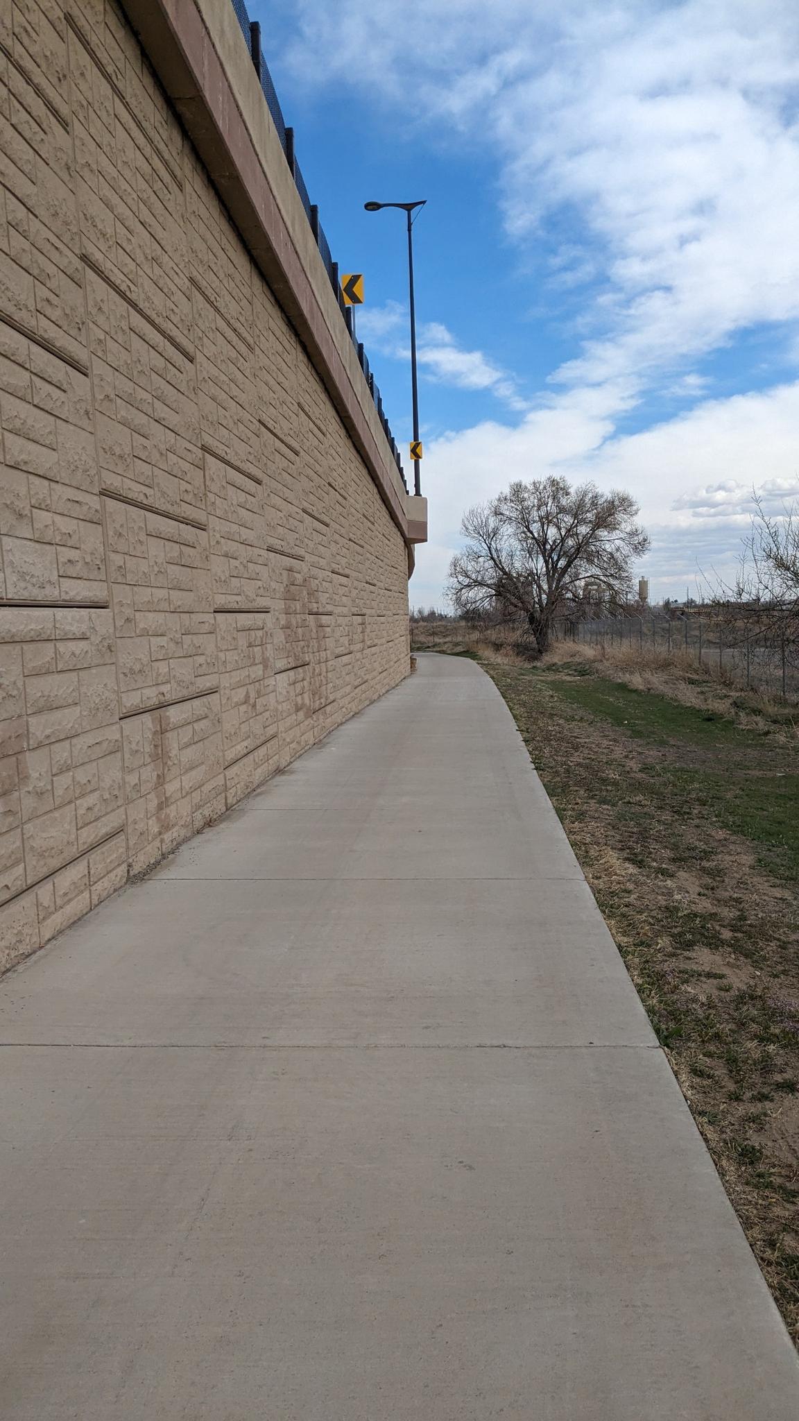 A concrete sidewalk next to a textured wall under a bright cloudy sky.