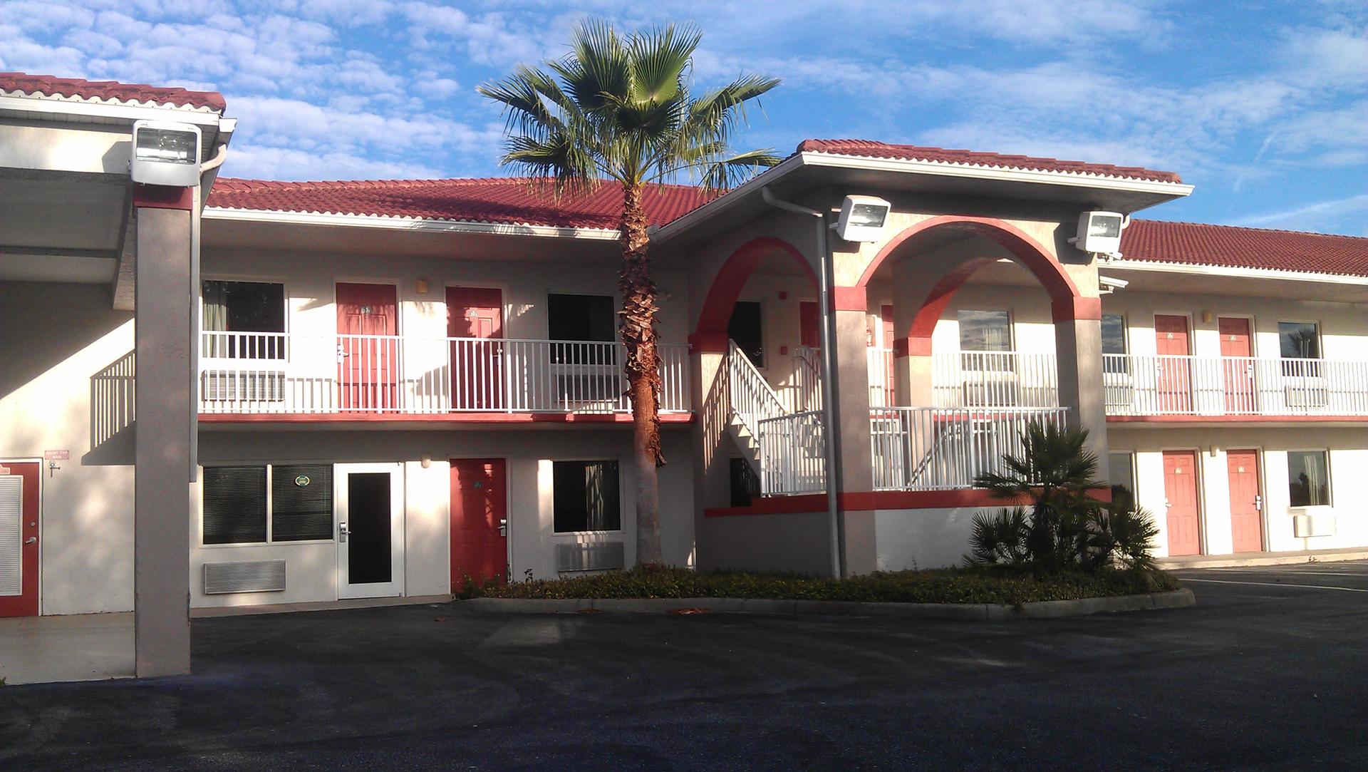 Sunny view of a two-story building with red roof, white walls, and palm tree.