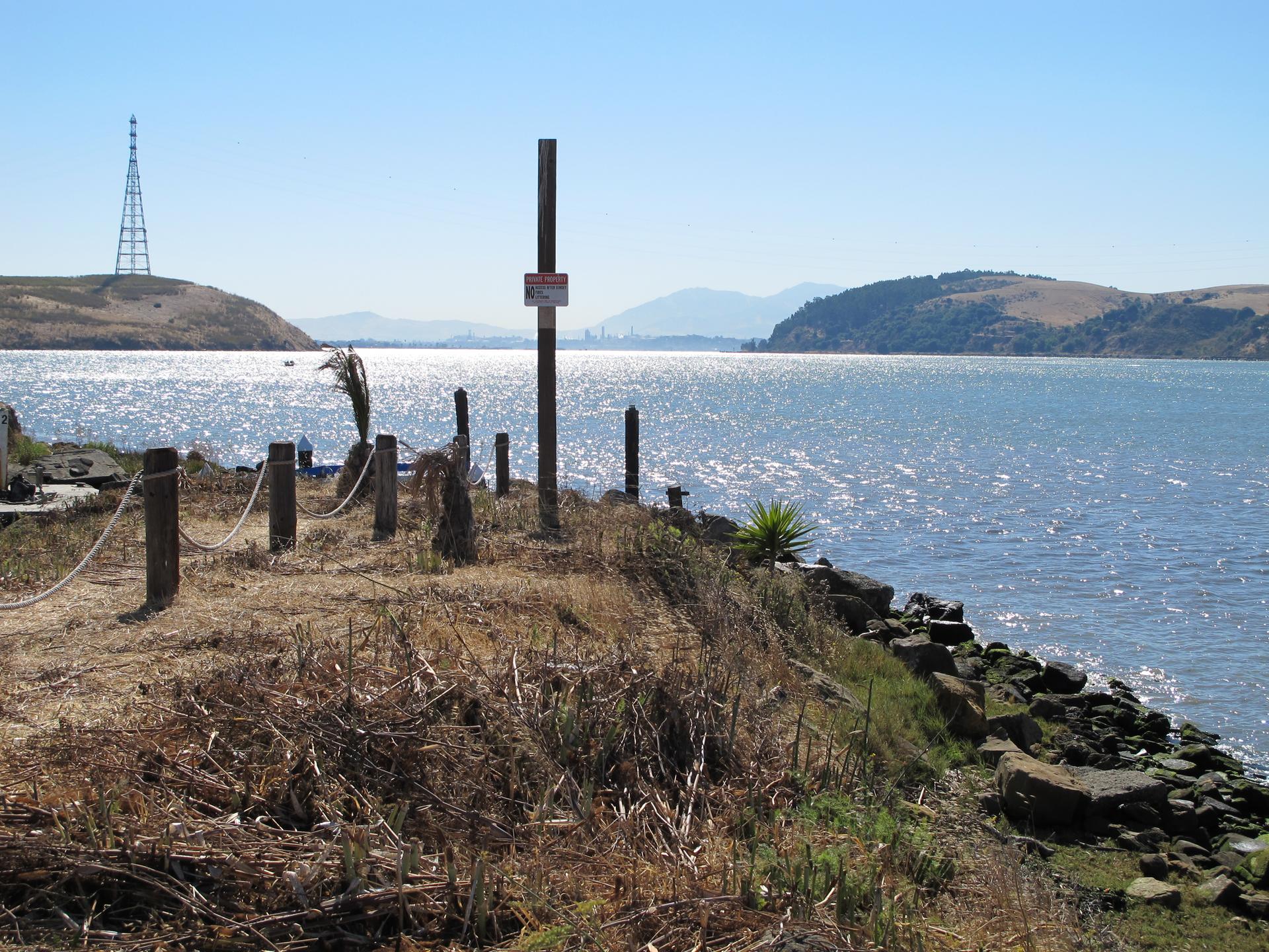 Rocky shoreline with wooden posts, distant tower, and sparkling water on a sunny day.