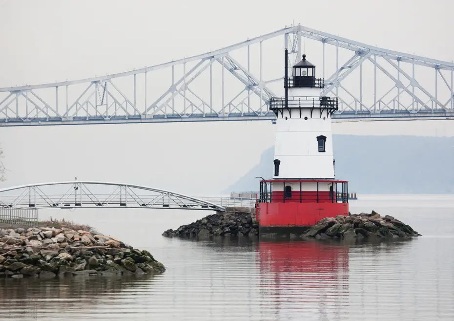 Red and white lighthouse in water with a large bridge behind it.