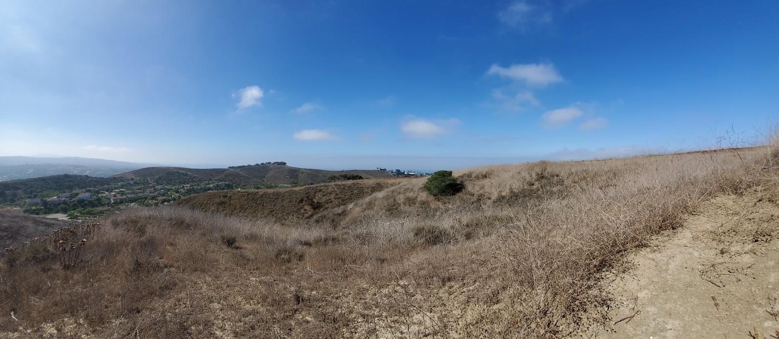 Panoramic view of dry, rolling hills under a bright blue sky with scattered clouds.