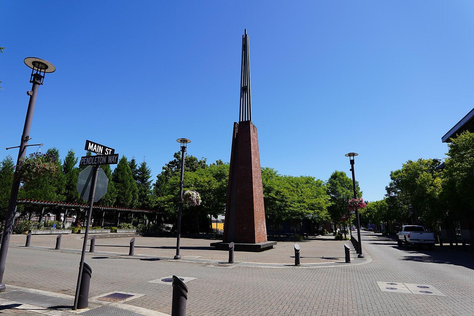 Tall brown monument in a paved town square under a clear blue sky.