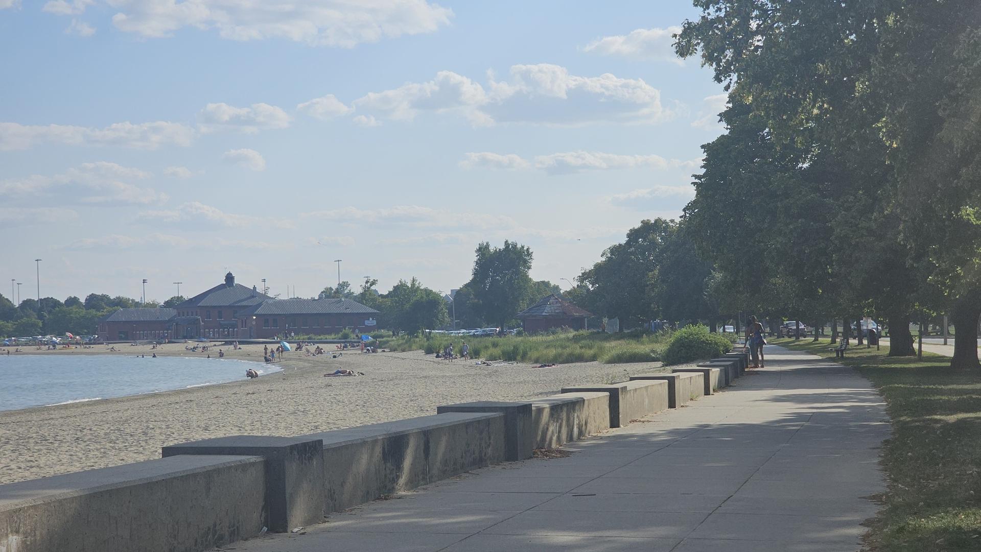 Sandy beach with people, paved path, and trees under a bright, cloudy sky.