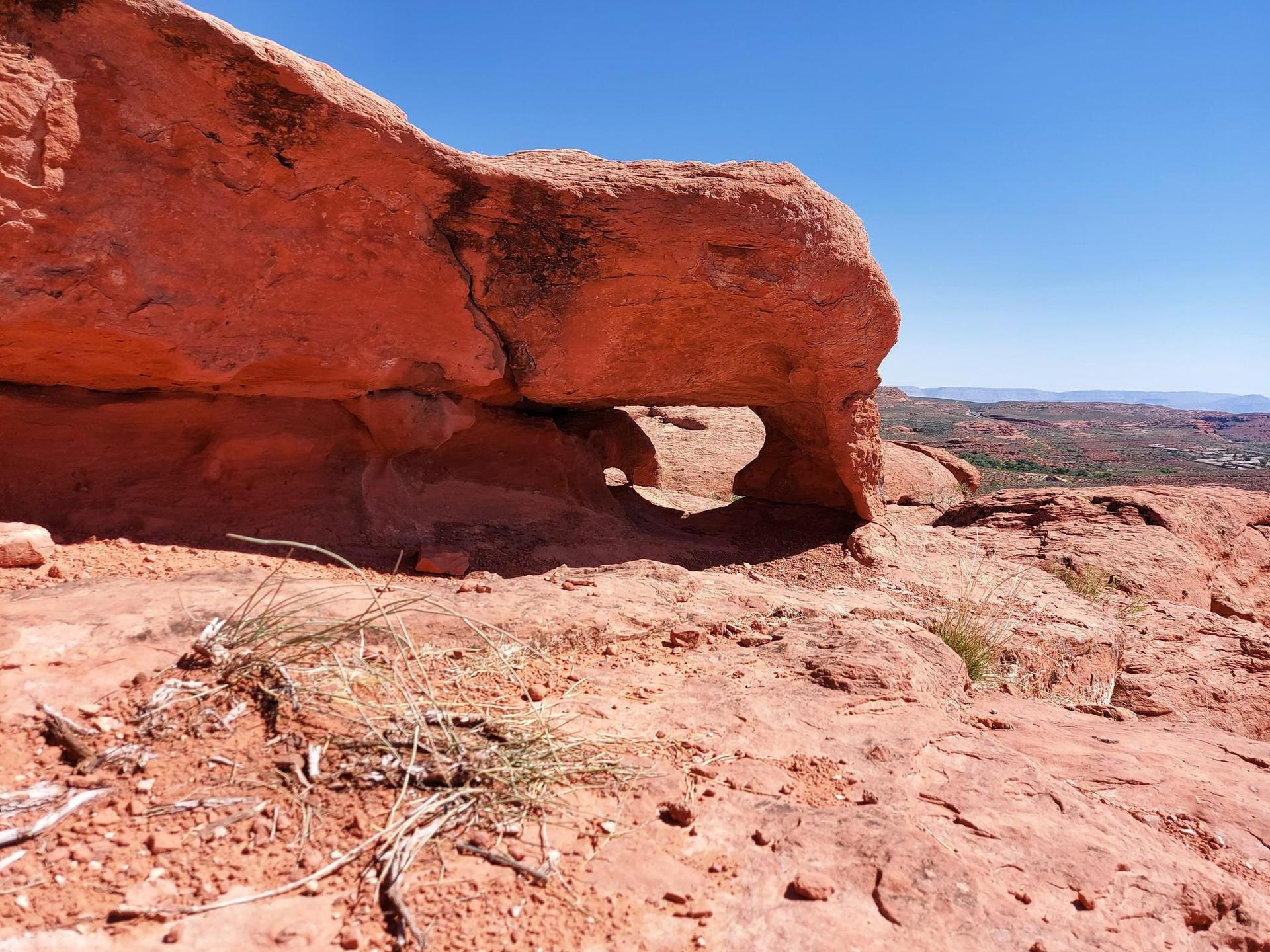Sunny desert landscape featuring a large red rock formation with a natural arch.