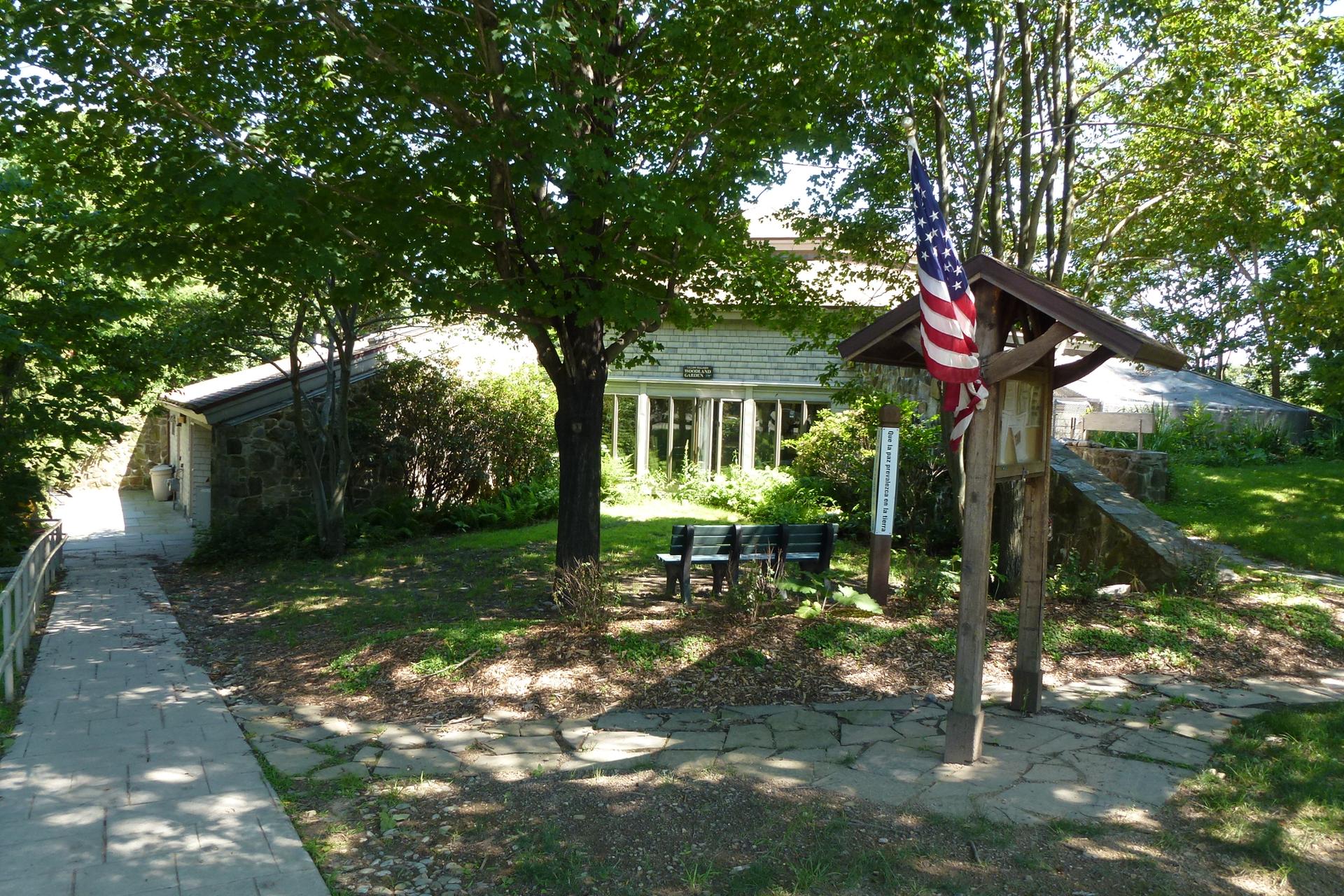 A building, flag, and path under dappled sunlight among green trees.