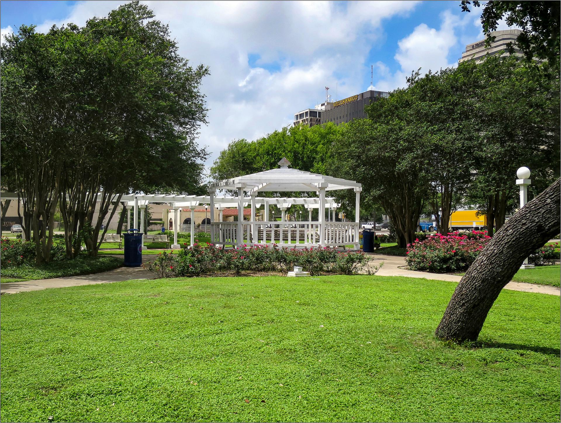 A white gazebo stands in a sunny park with green grass and trees.