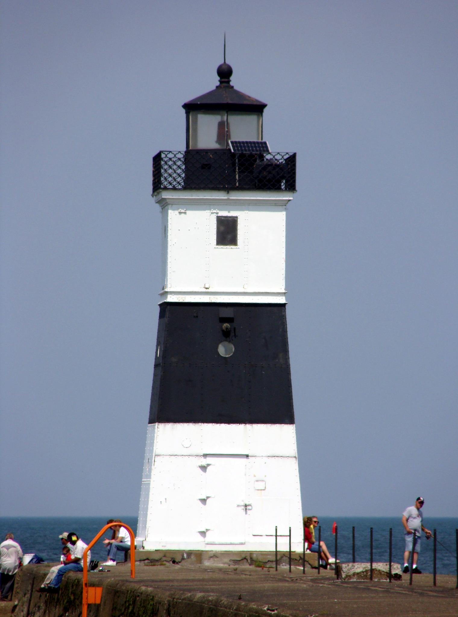 A black and white lighthouse stands tall with people nearby on a sunny day.