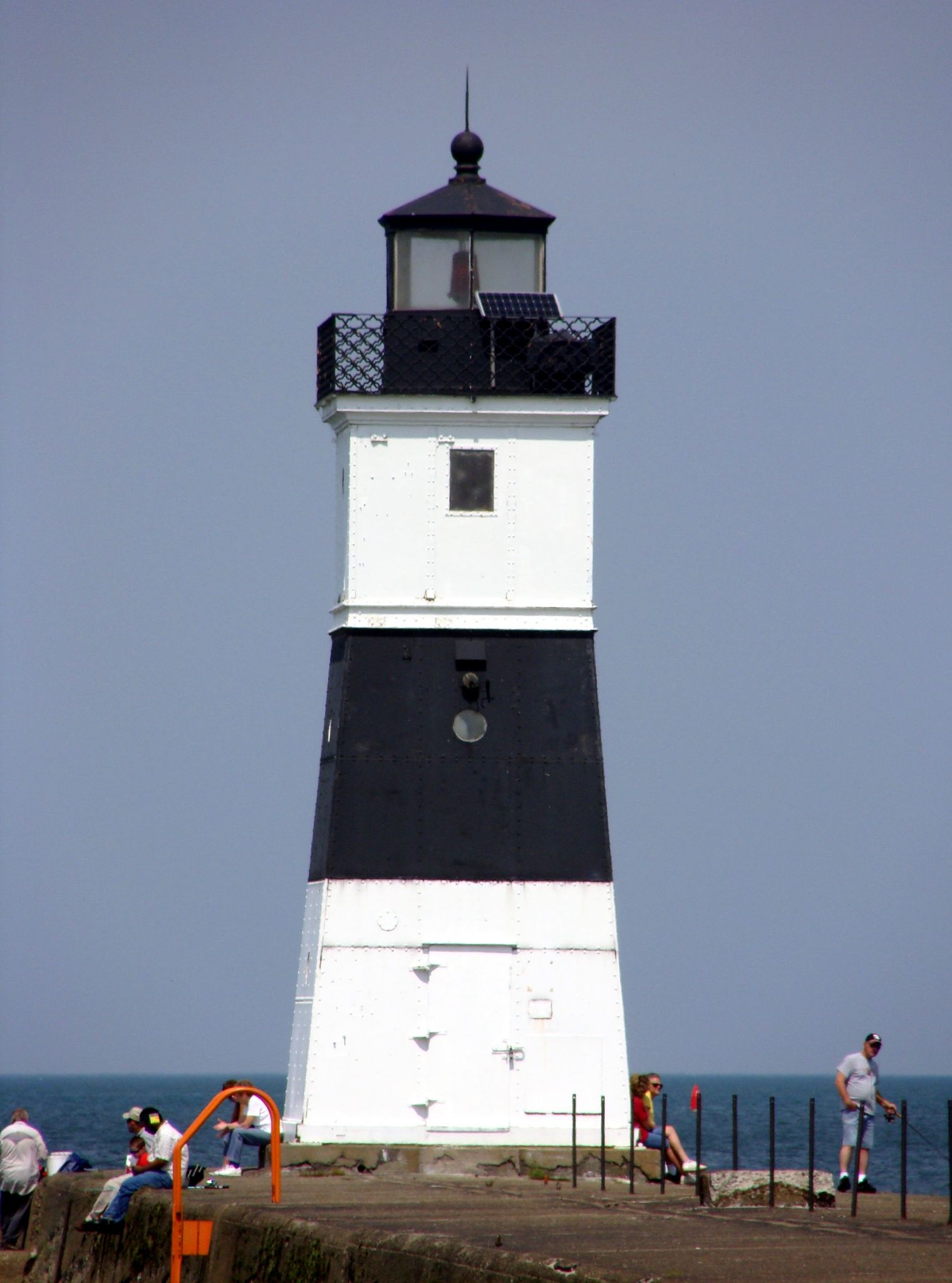 A black and white lighthouse stands tall with people nearby on a sunny day. - North Pier Trail