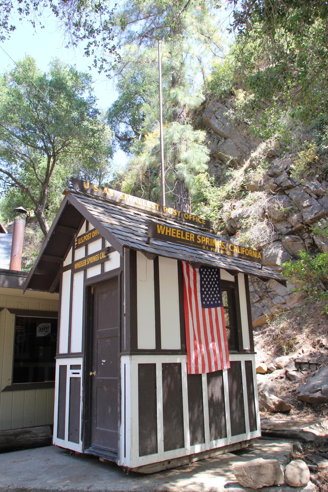 Small rustic building with an American flag, nestled against a rocky, wooded hillside.