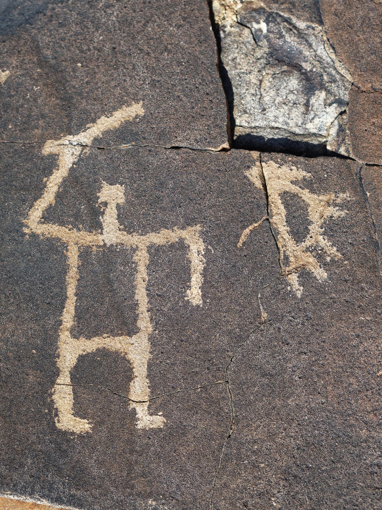 Close-up of ancient petroglyphs carved into dark rock, showing human-like figures.