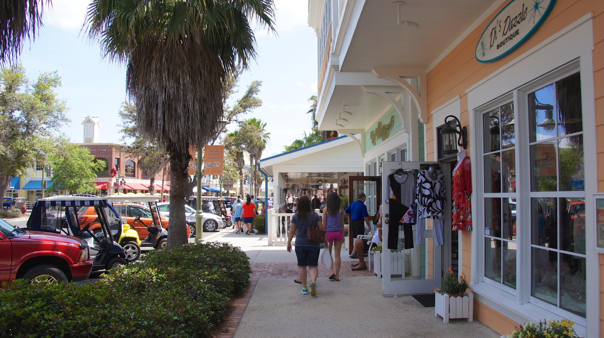 People stroll past shops and palm trees on a bright, sunny day.