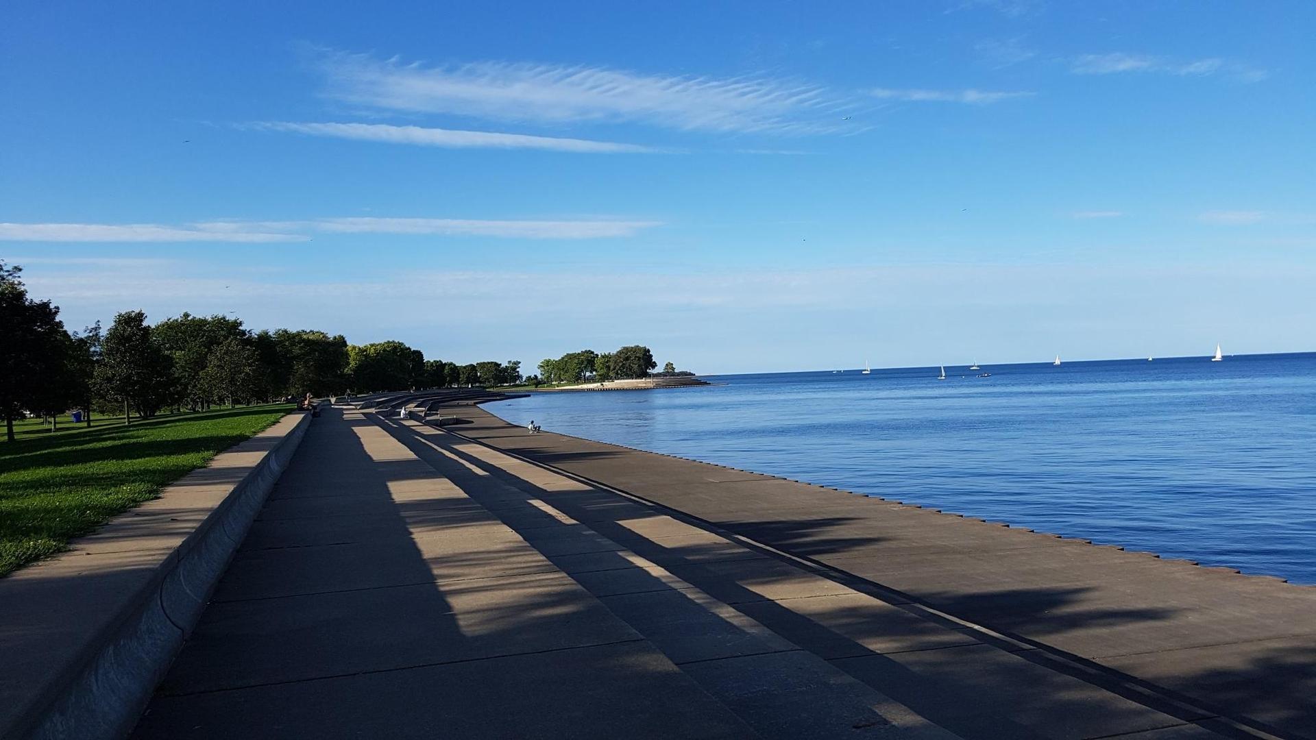Sunny concrete path alongside a vast blue lake with distant sailboats.