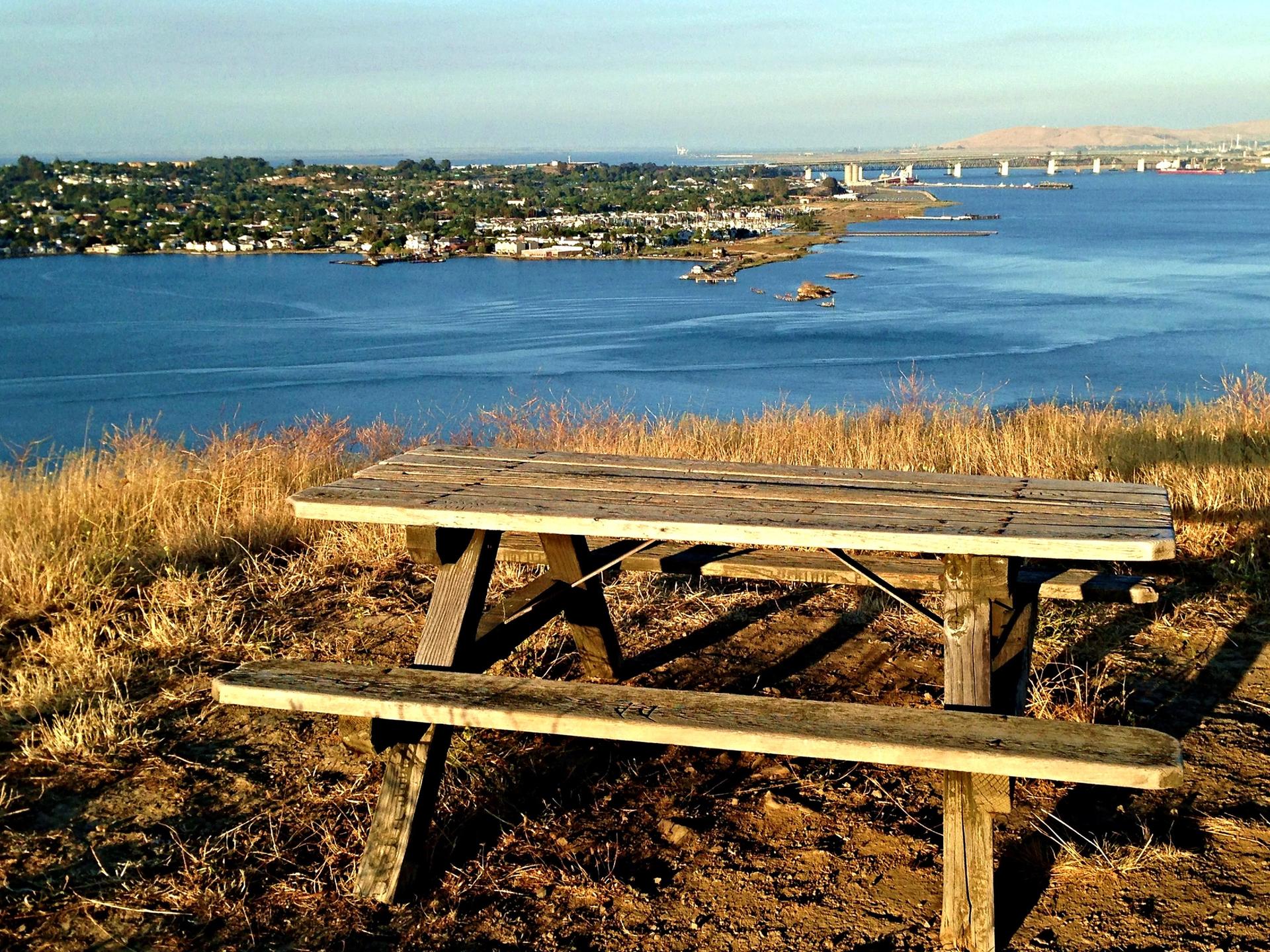 A wooden picnic table overlooks a vast bay and distant town from a hill.