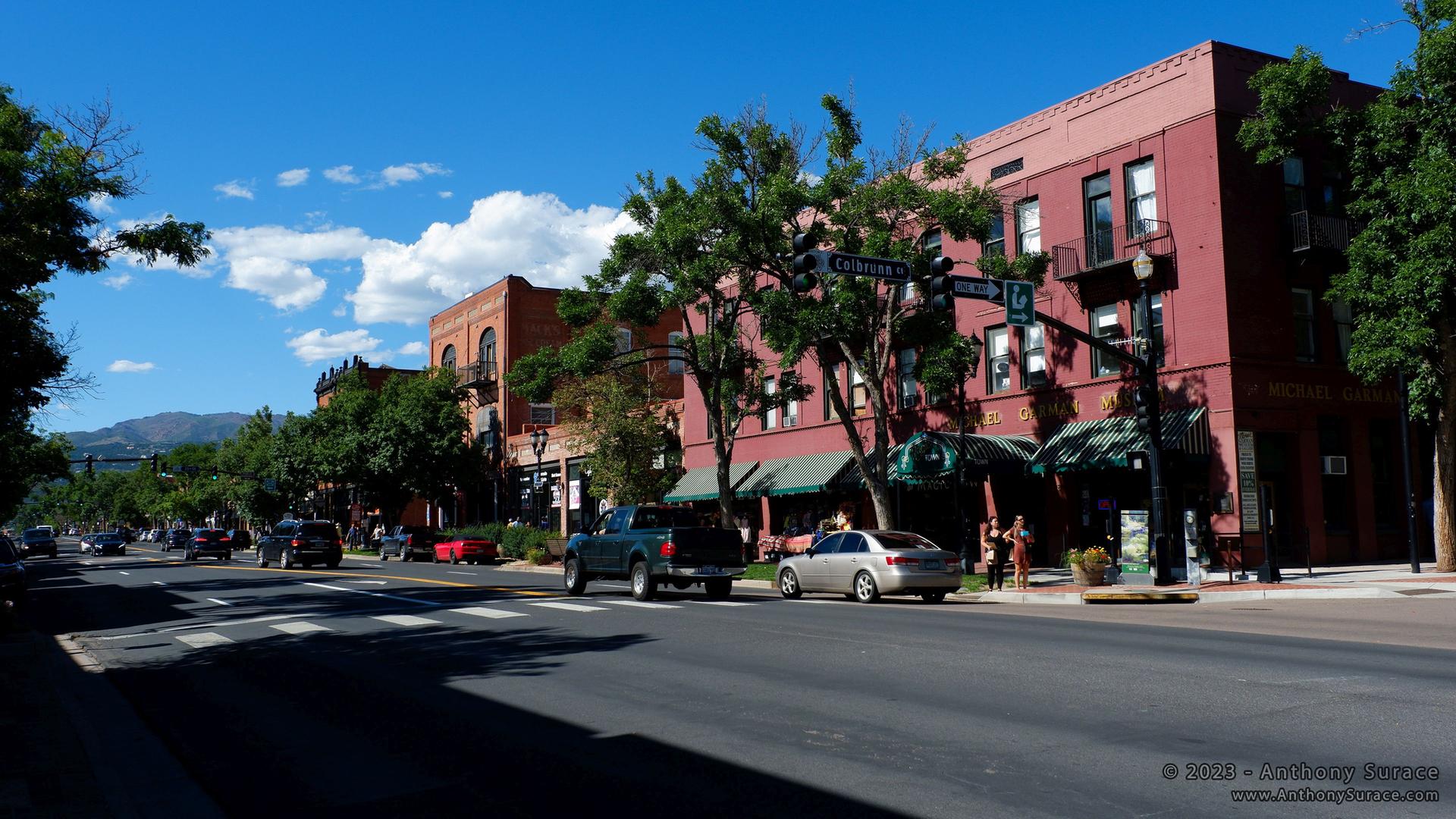 Sunny street with brick buildings, parked cars, and people walking on the sidewalk.