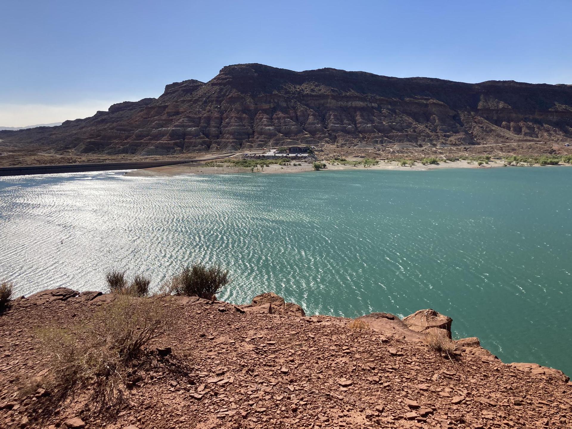 Turquoise water reflects sunlight, with arid mountains and rocky foreground under clear sky.