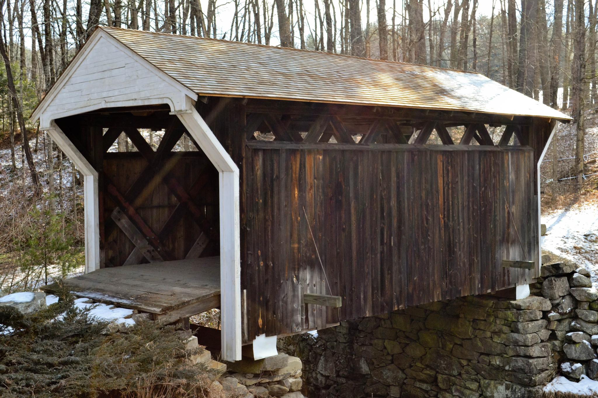 A rustic wooden covered bridge stands over snowy ground with bare trees.