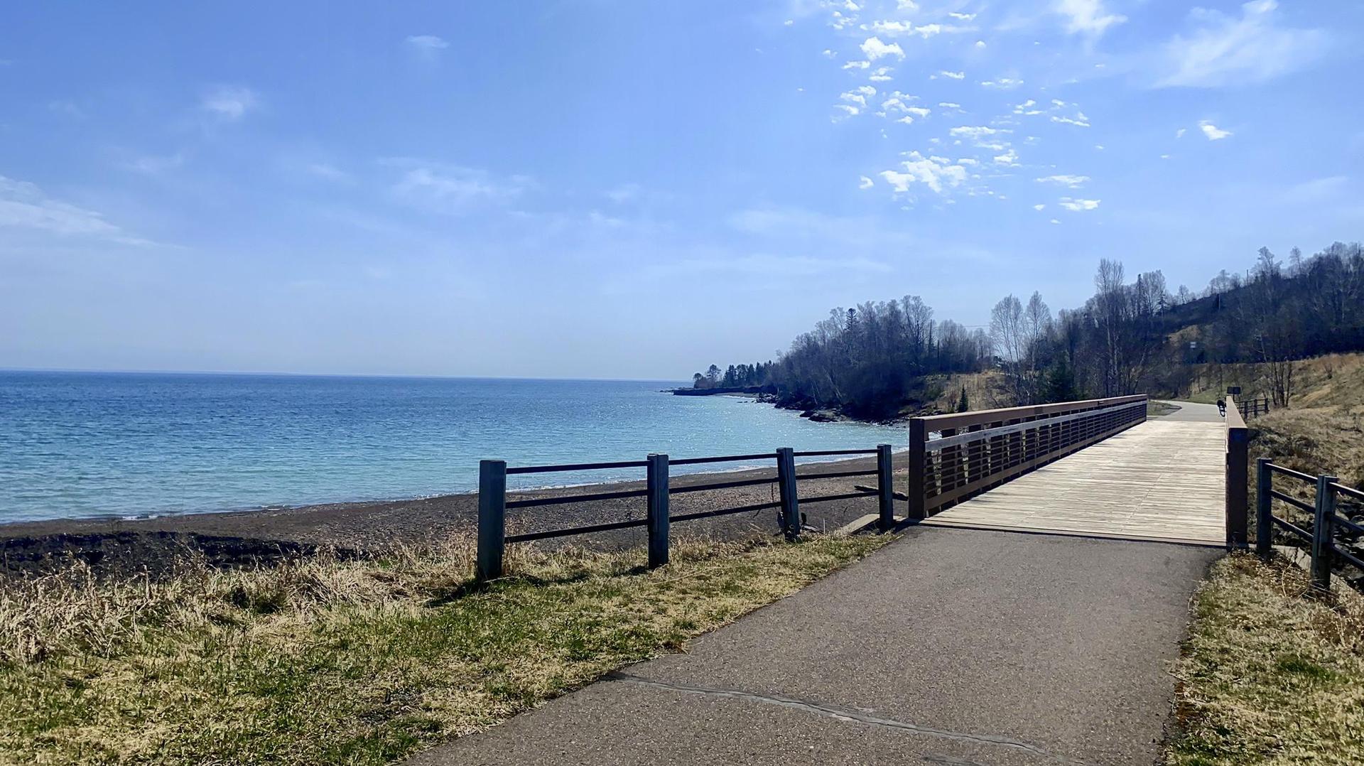 Paved path and wooden bridge beside blue water, grassy bank, and distant trees under a bright sky. A person walks in the distance.