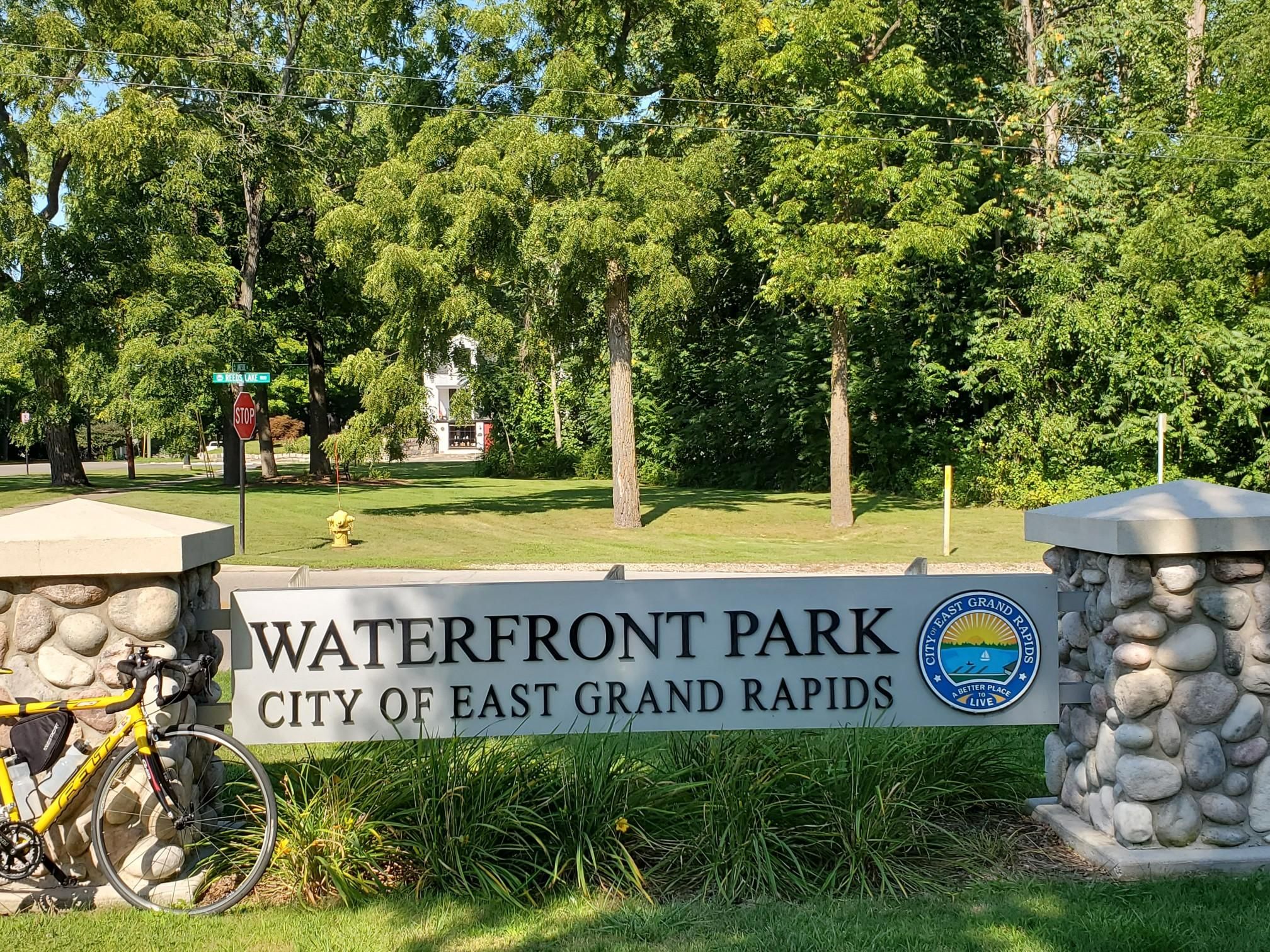 A yellow bicycle parked beside a stone park sign under bright sunlight. - Reeds Lake Dock – Waterfront Park loop from Wealthy Theater