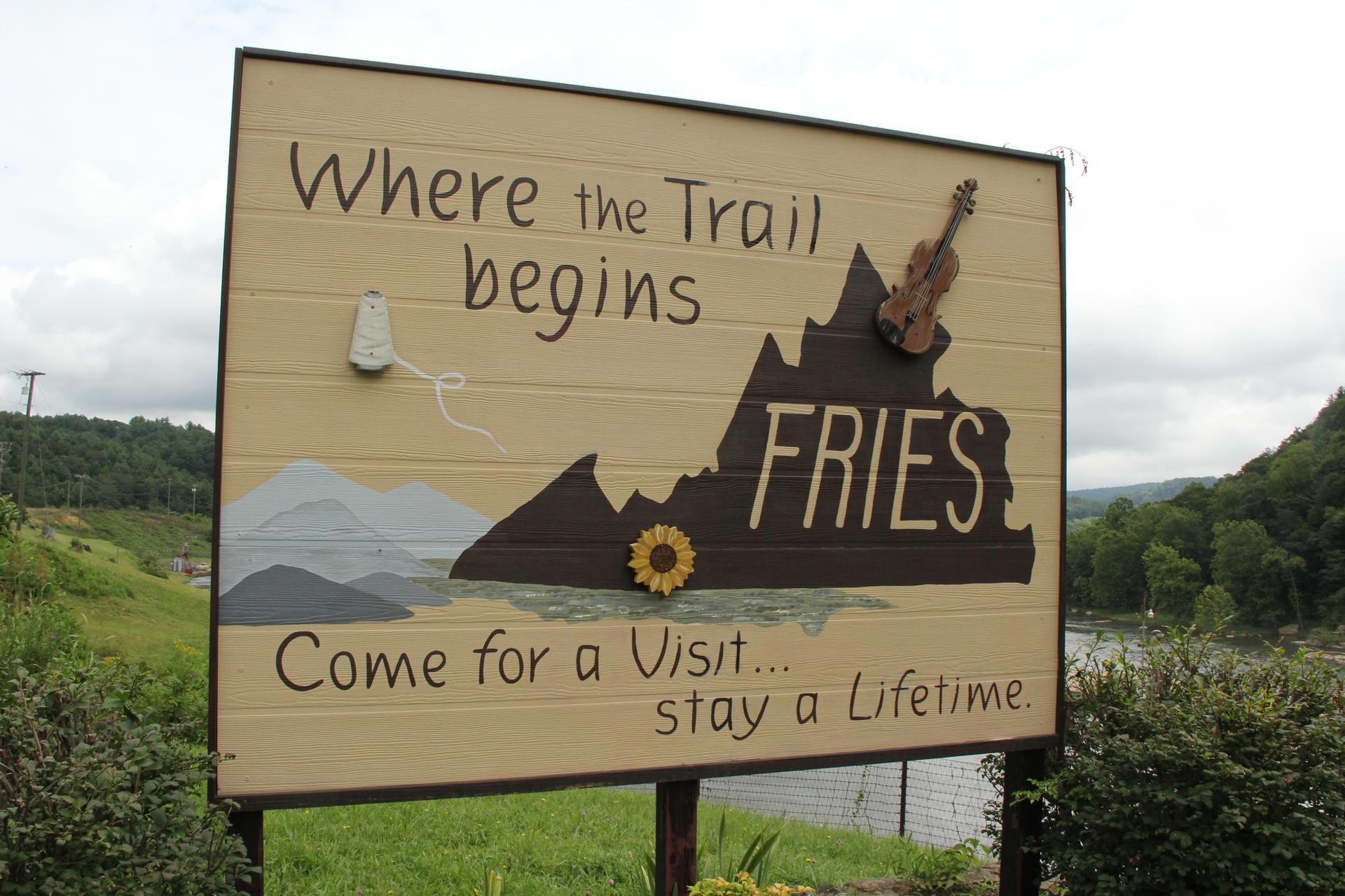Large wooden sign with text, mountains, and a violin under a cloudy sky.