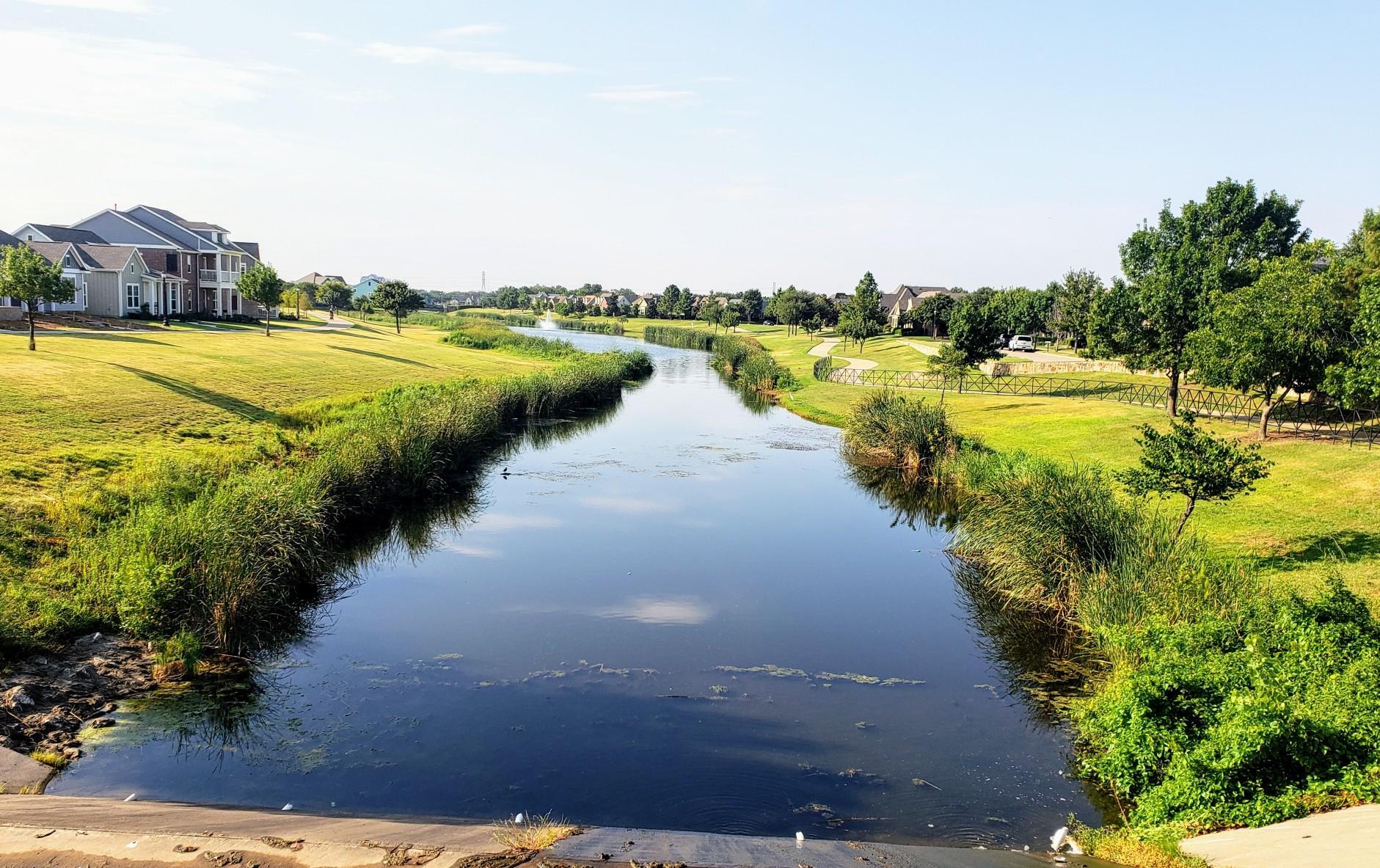 A calm river flows through green banks with houses under a bright sky.
