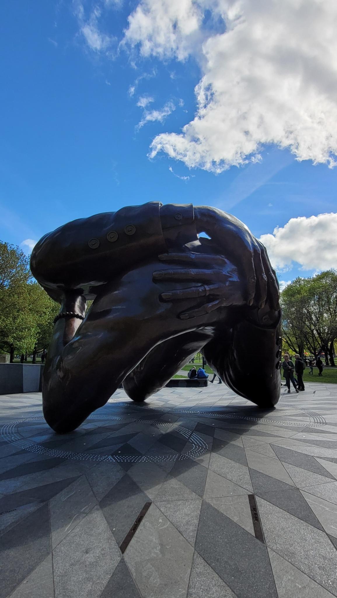 Large dark sculpture of intertwined hands under a bright blue sky.