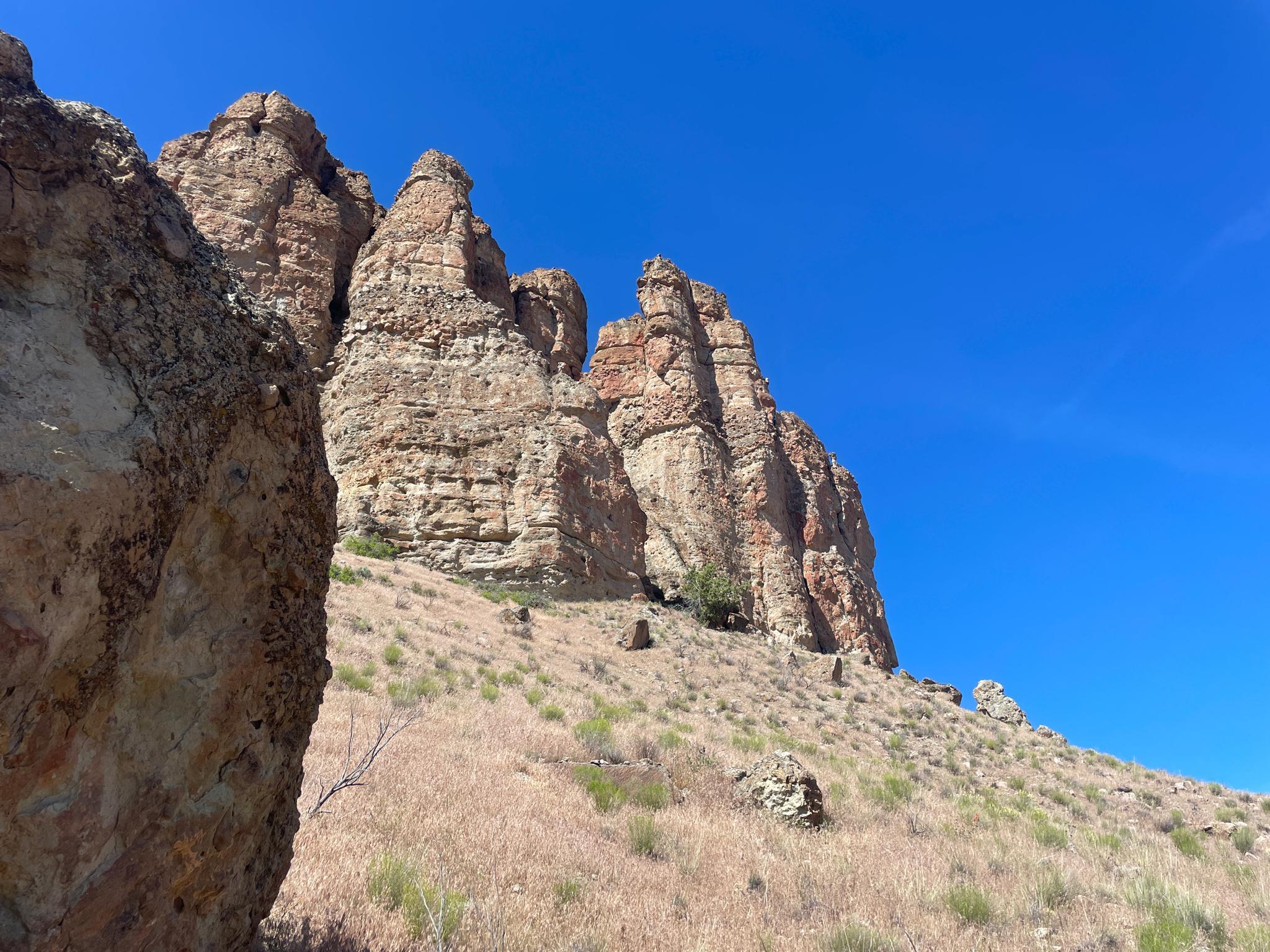 Large, weathered rock pillars rise from a dry, grassy hillside under blue sky.