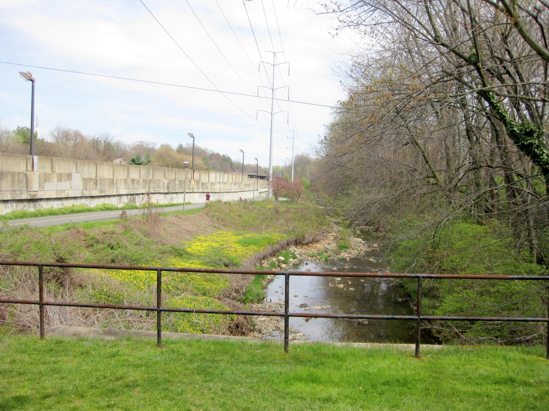 Stream with green banks, paved path, power lines, and distant wall.