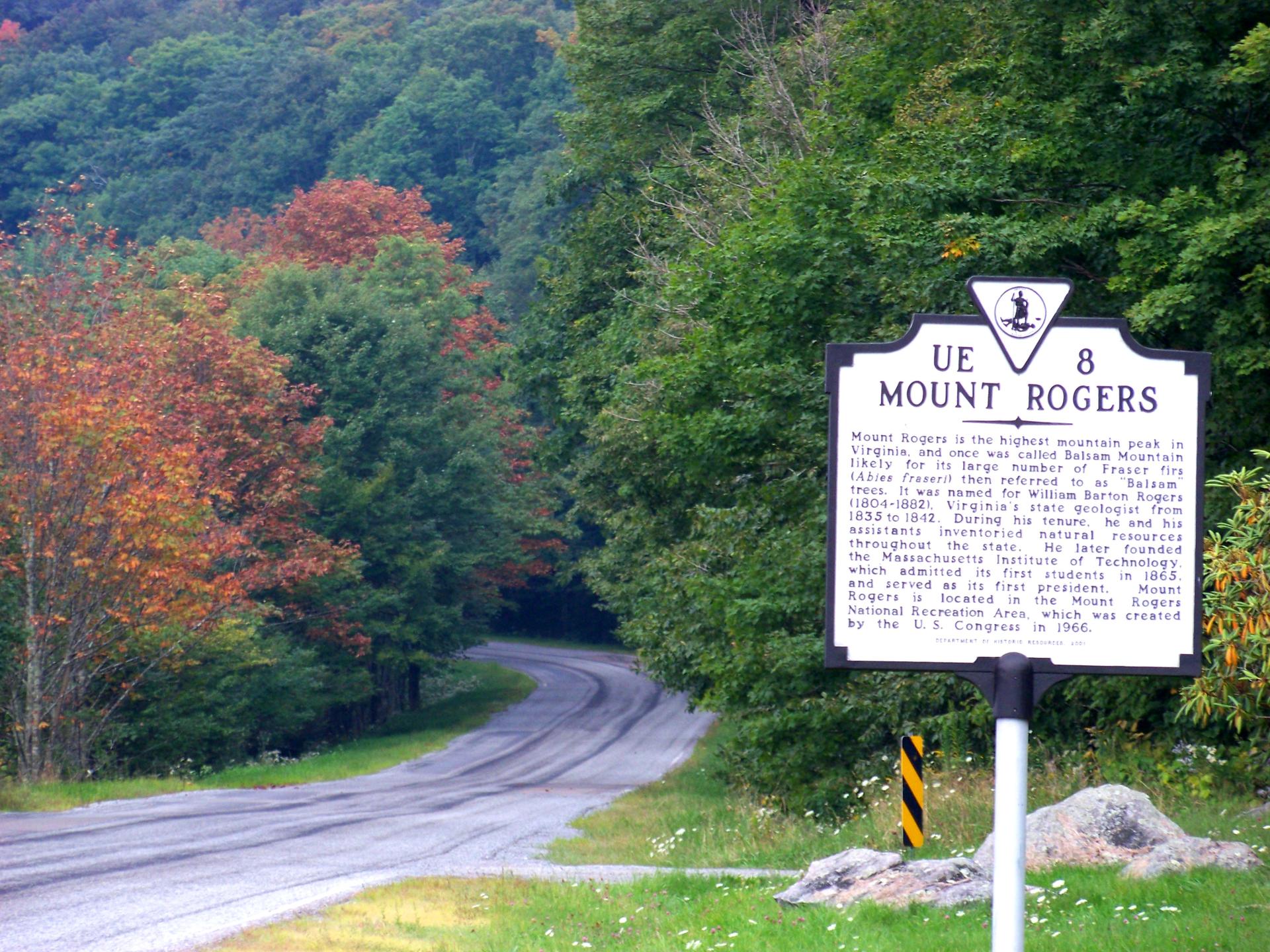 A white roadside sign stands beside a winding road through autumn trees.