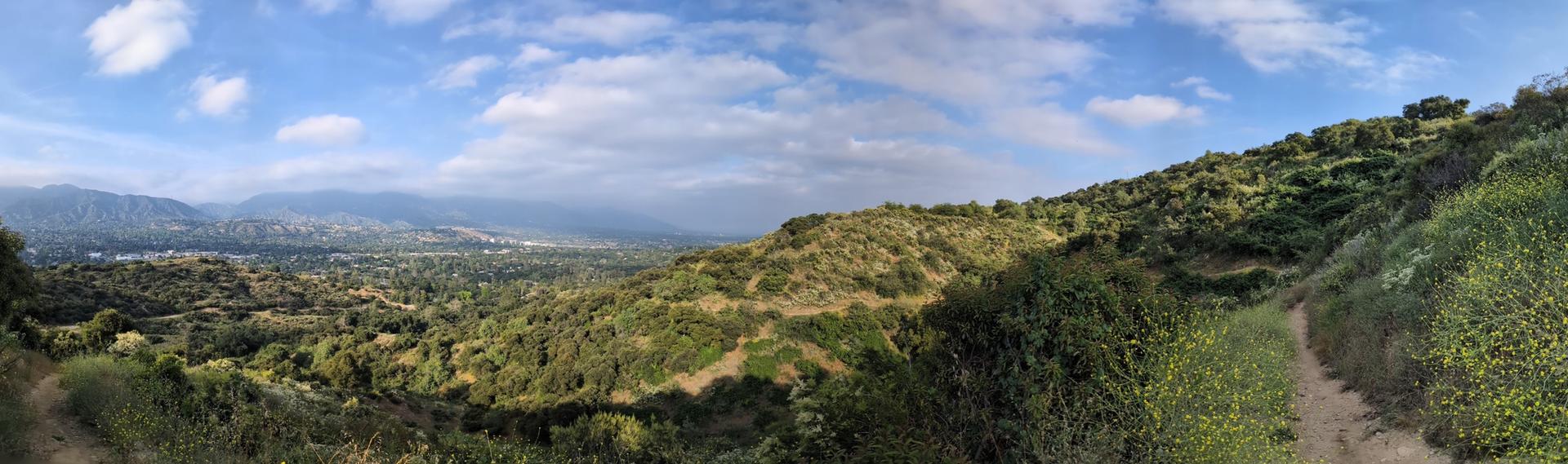 Panoramic view of a green valley with distant city and mountains under cloudy sky.
