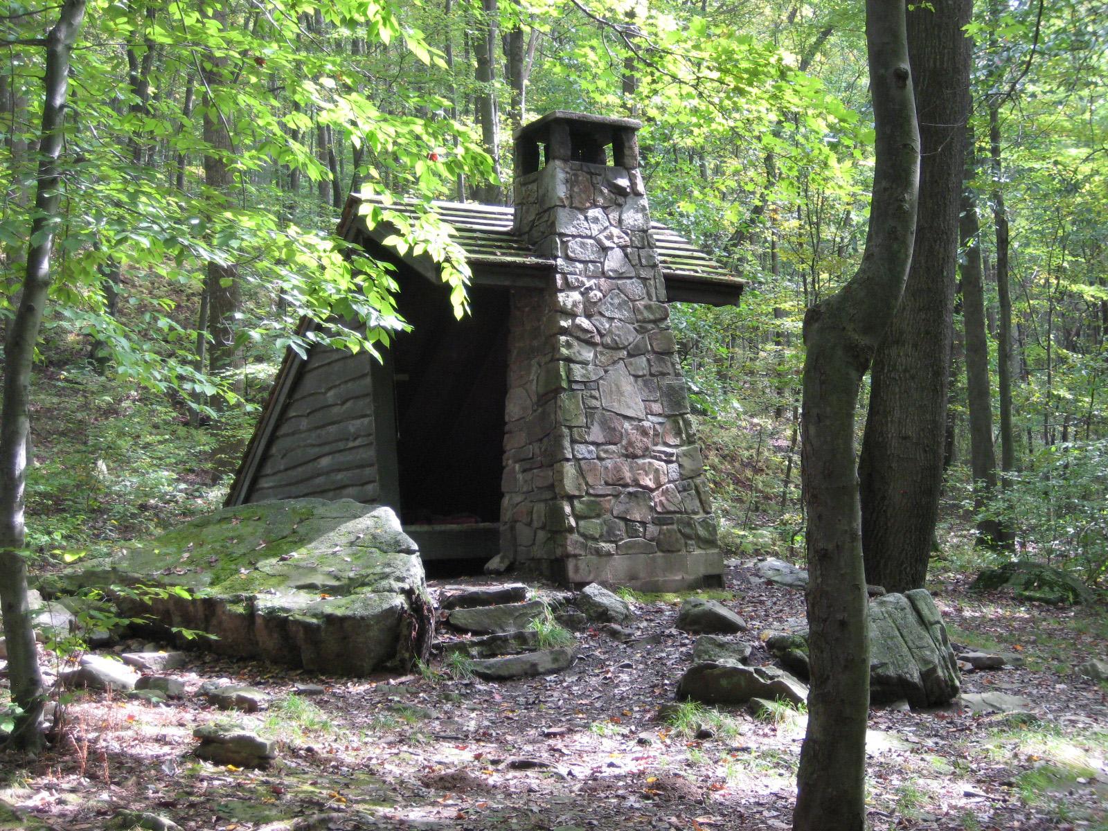 A rustic stone and wood shelter with a large chimney in a green forest.
