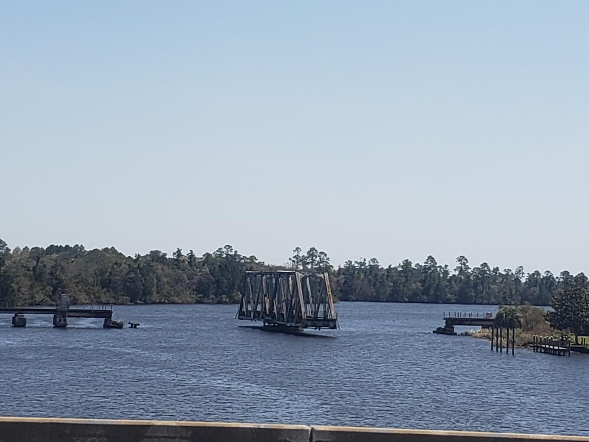 Metal bridge structure over wide water, with distant forested banks under clear sky.