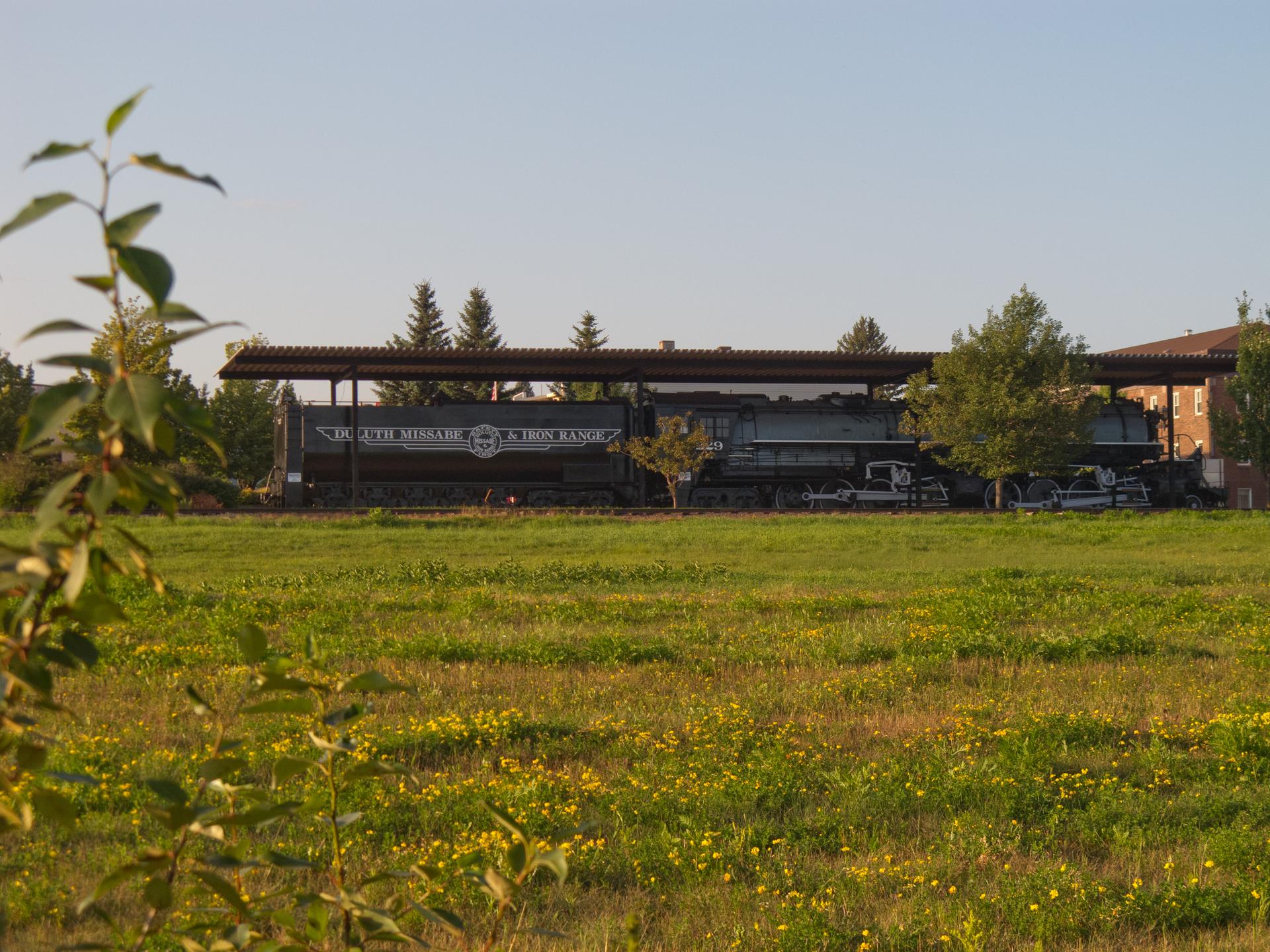 An old train sits under a shelter in a sunny, grassy field.