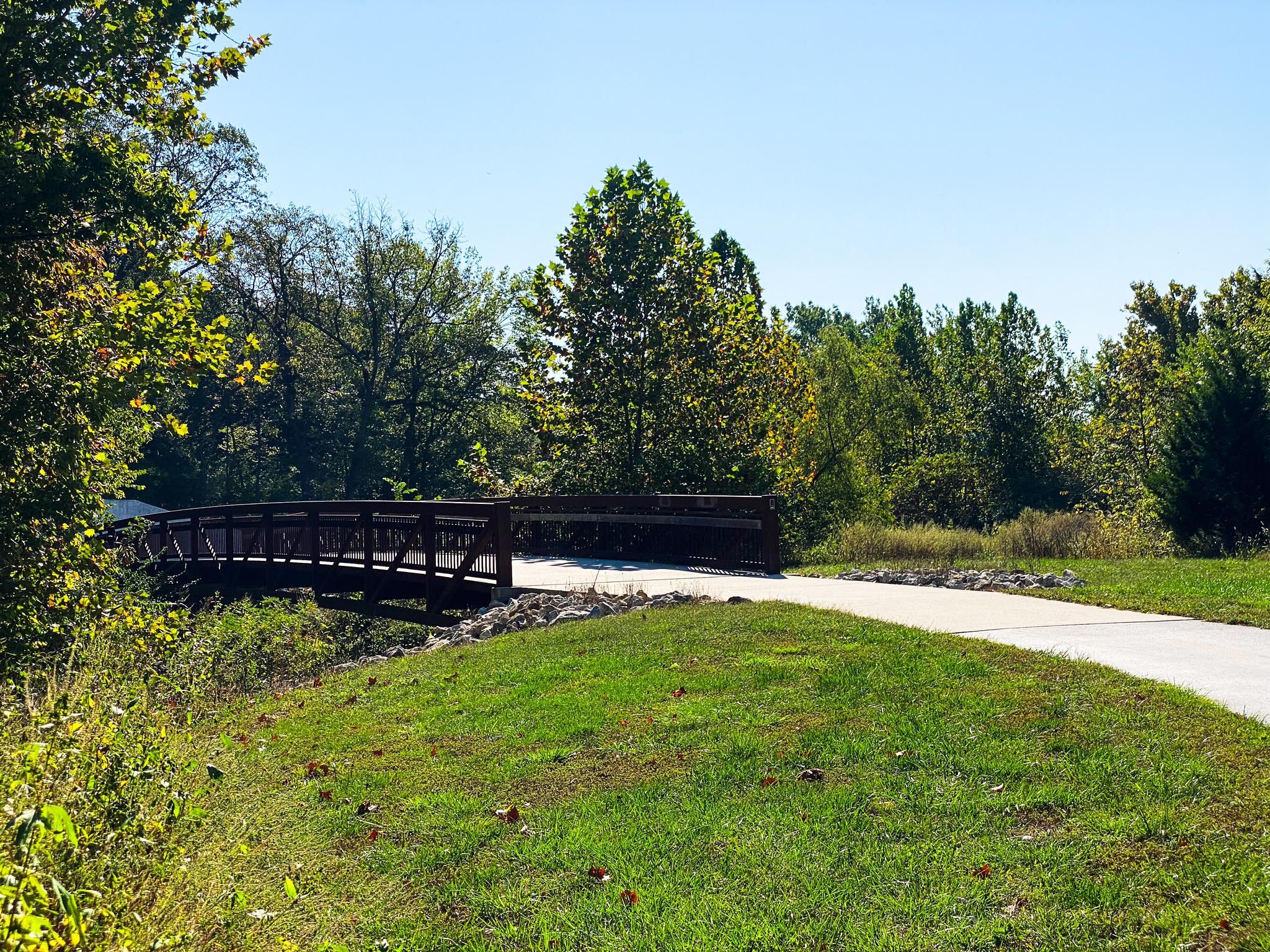 Sunny view of a wooden bridge and paved path through green trees. - Arthur & Betty Verhaeghe Park – Lake Olathe loop from RideKC
