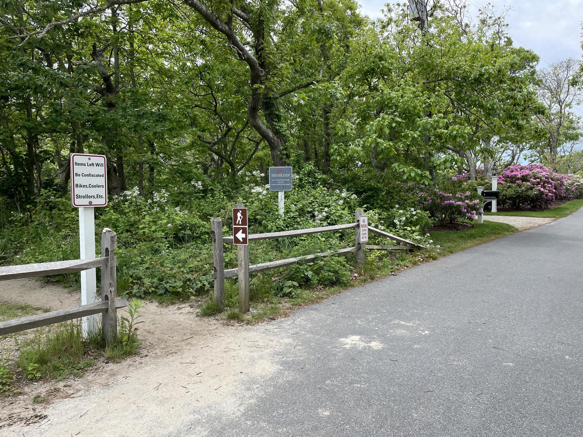 Wooden fence and signs mark a trail entrance next to an asphalt road.