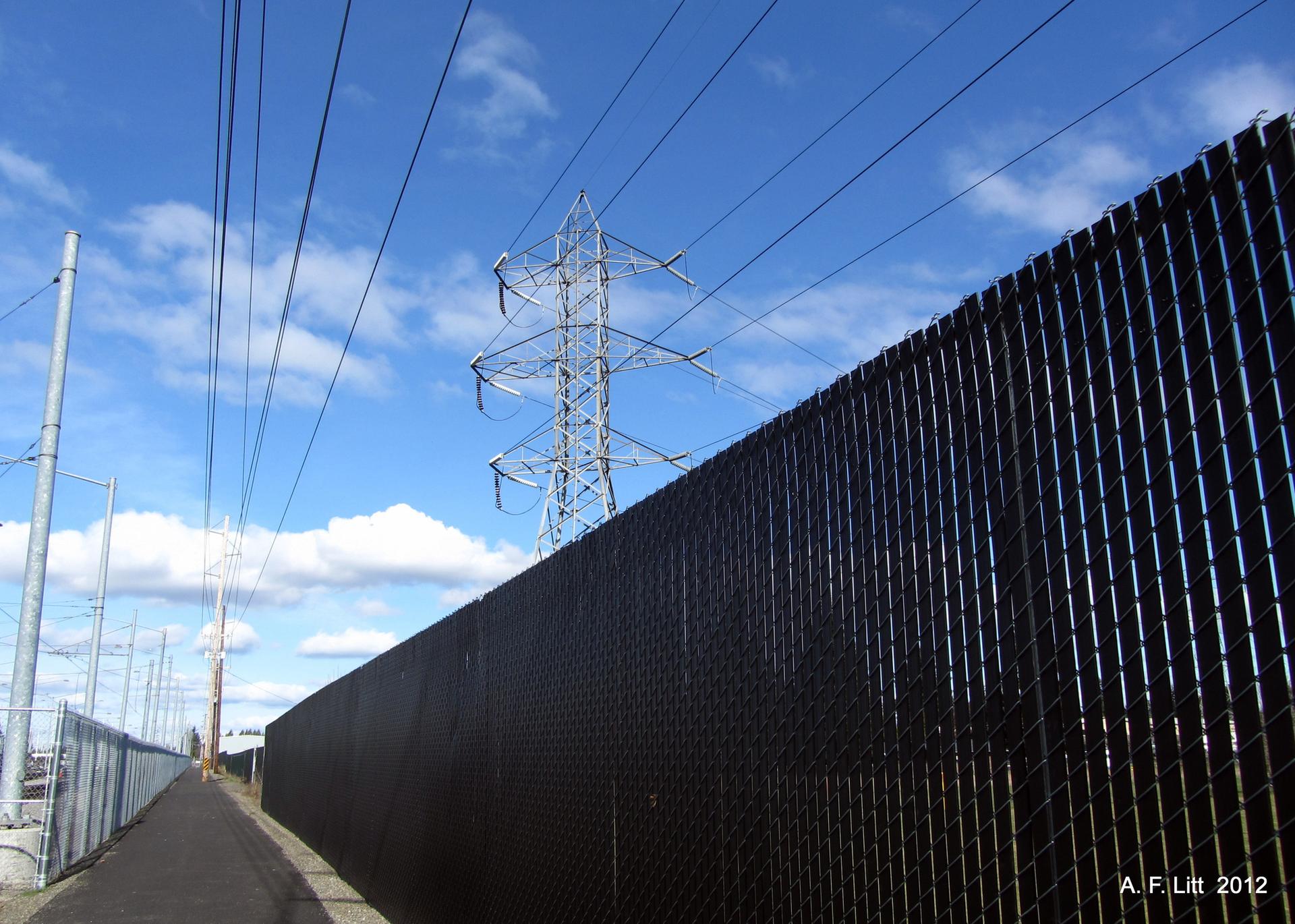 Power lines and a transmission tower rise above a black fence and paved path.