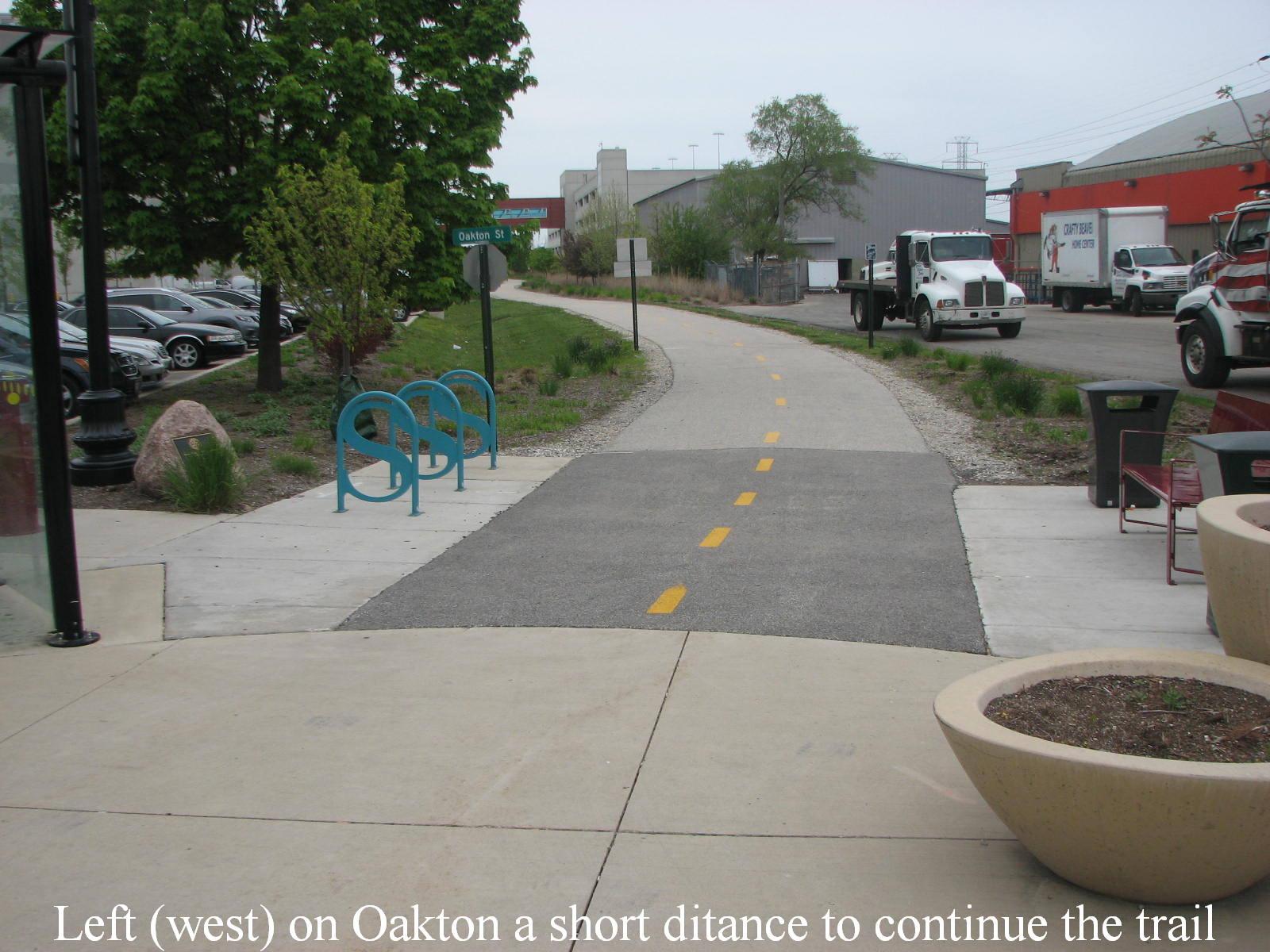 Paved urban path with bike racks, parked cars, and industrial buildings under overcast sky.