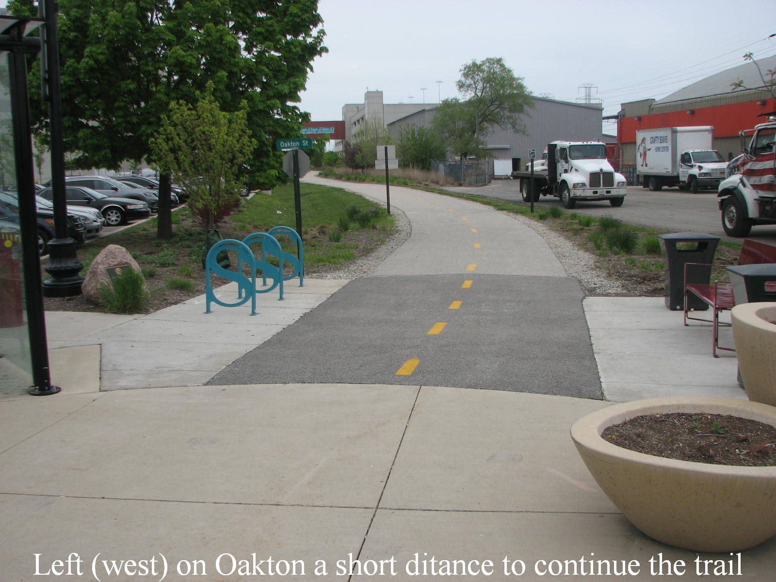 Paved urban path with bike racks, parked cars, and industrial buildings under overcast sky. - Valley Line Trail