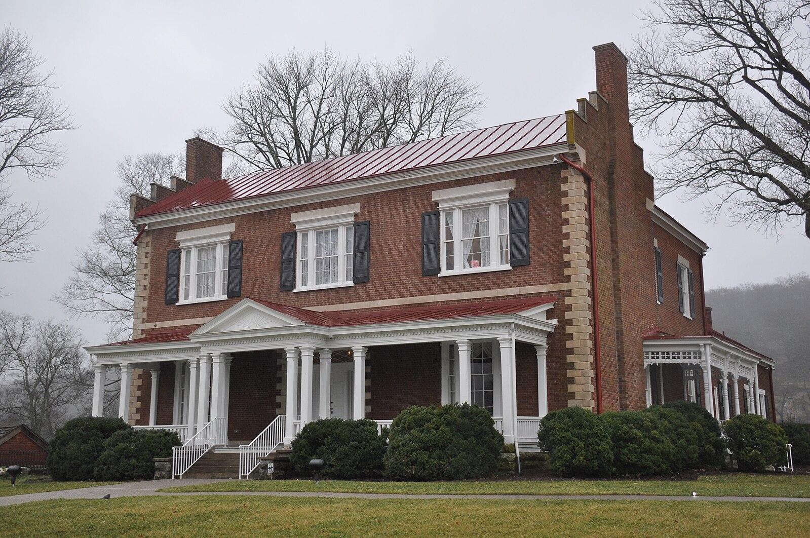 Large brick house with red roof, white columns, and bare trees on a cloudy day.