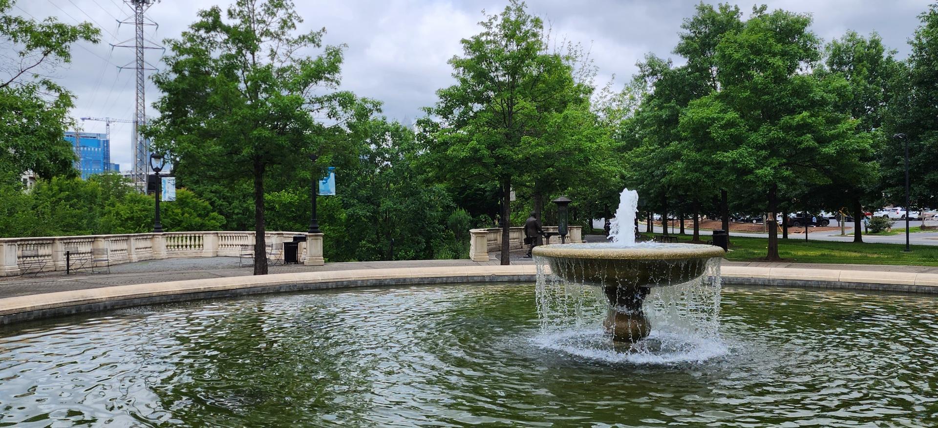 A large fountain in a circular pool under an overcast sky, surrounded by trees.