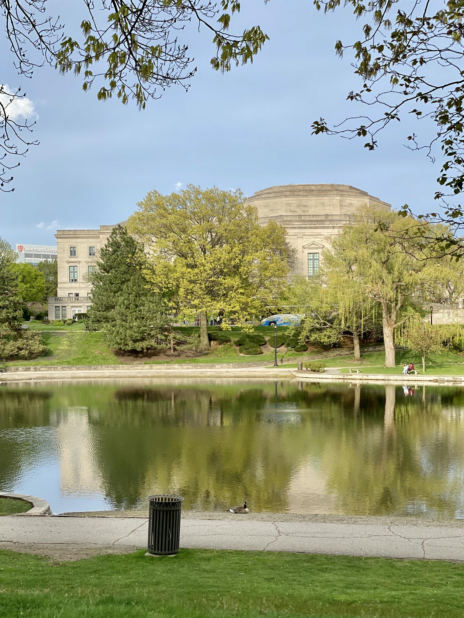 Large building reflected in a calm lake with green trees and distant people.