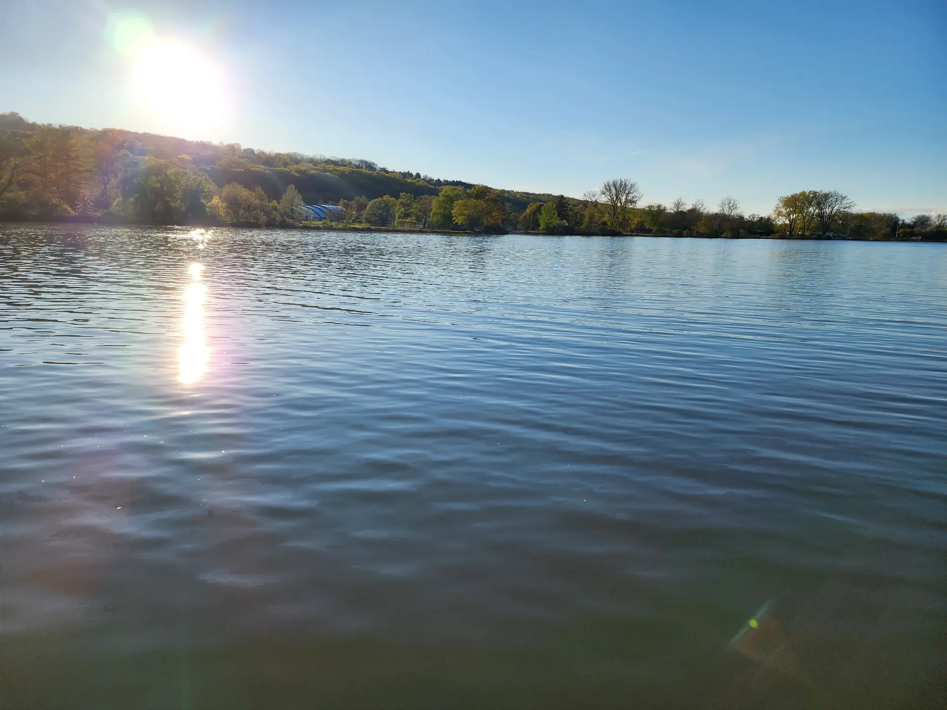 Bright sun reflects on rippling water with distant green hills under a clear sky.